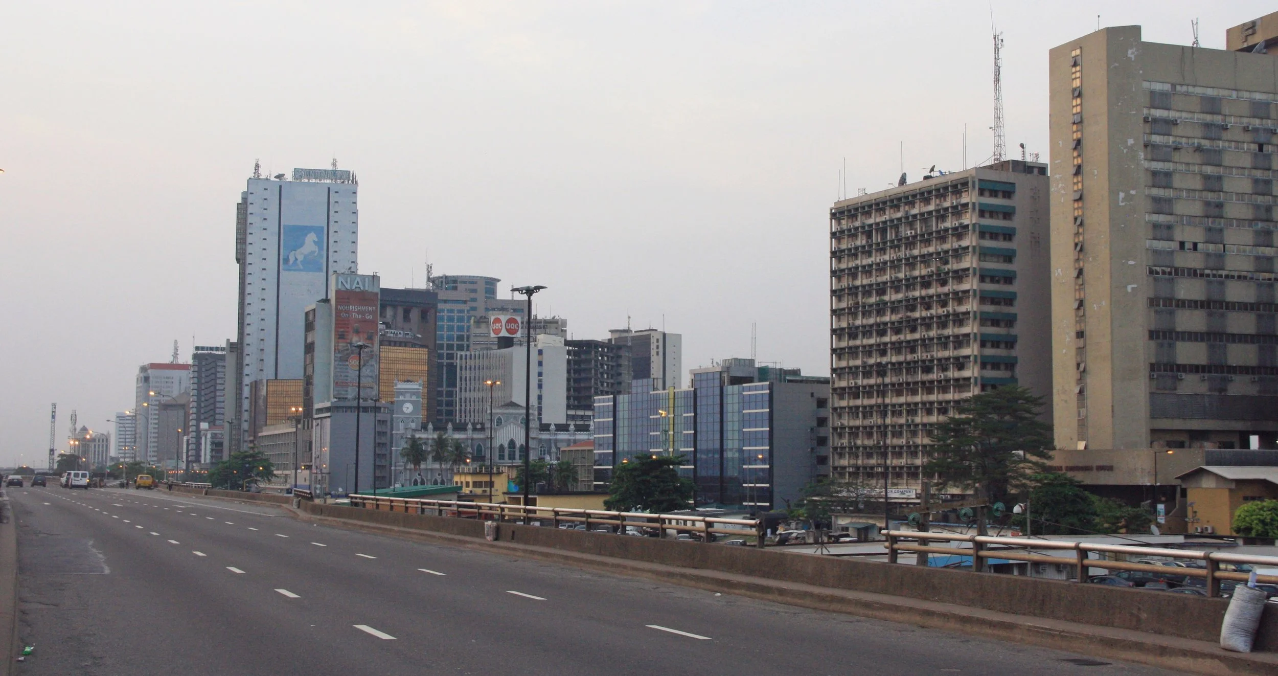 A city skyline with tall buildings, some with colorful signs and advertisements, viewed from a highway with moving vehicles and streetlights.