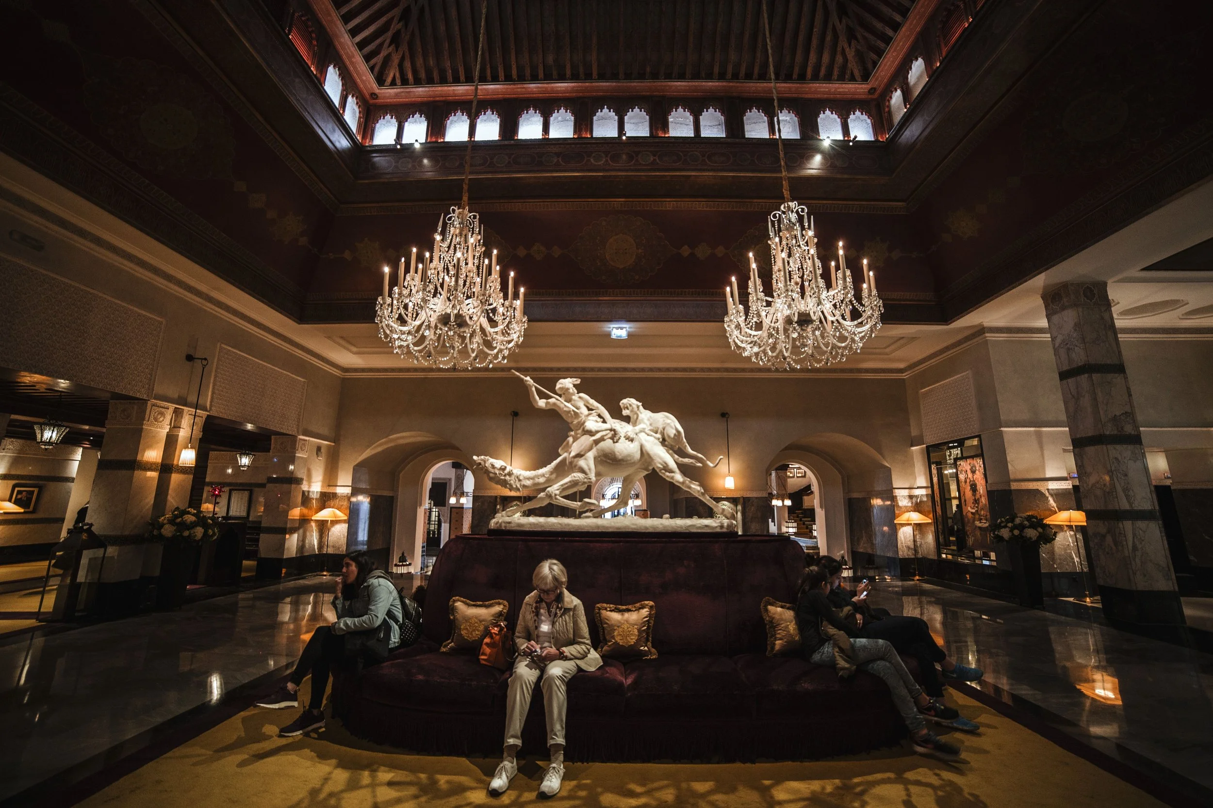 Interior of a grand hotel lobby with chandeliers, a marble sculpture of horses and a person sitting on a couch, with decorative walls and ceiling.