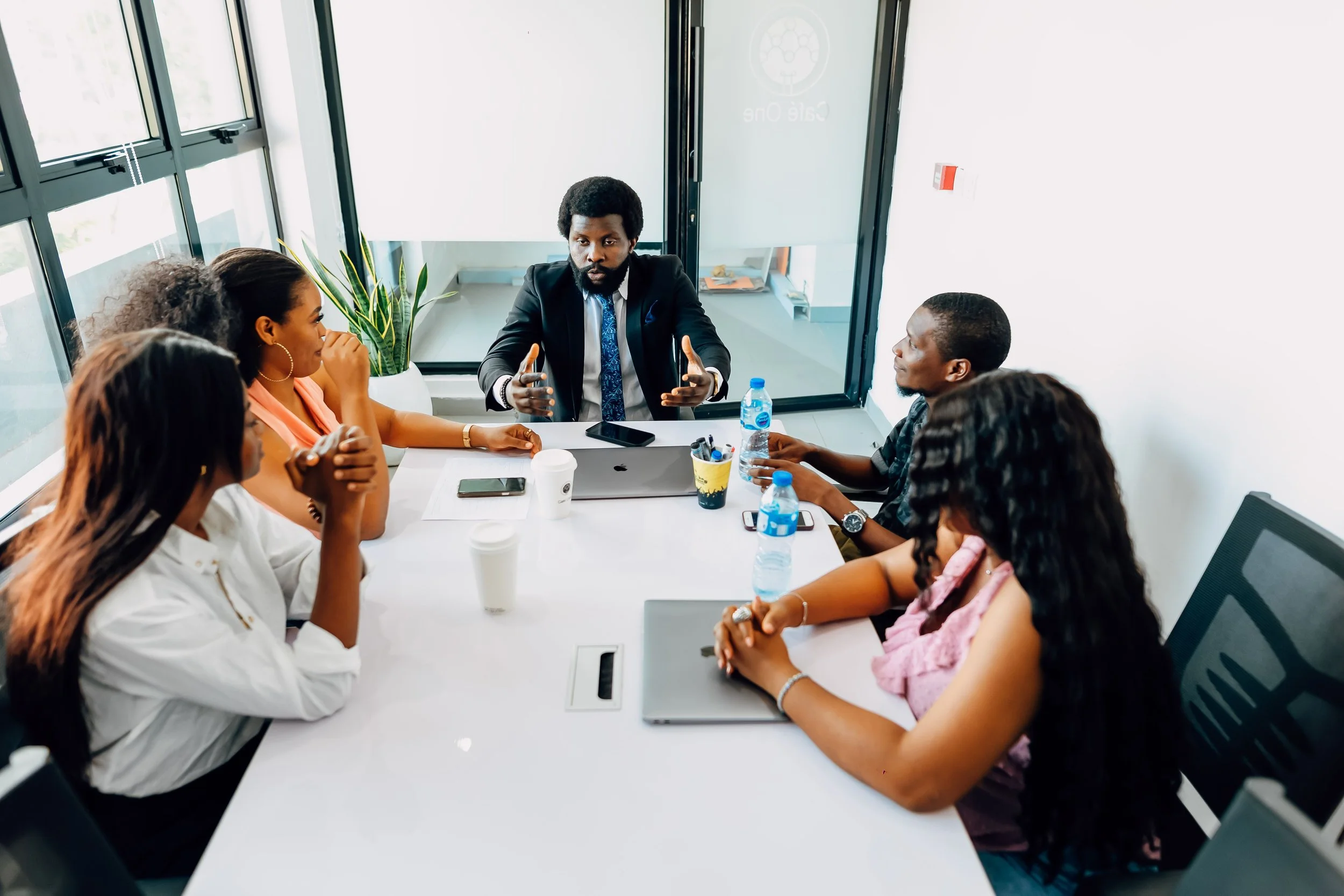 Five people sitting around a conference table engaged in a discussion, with a man in a suit speaking and others listening, in a modern office.