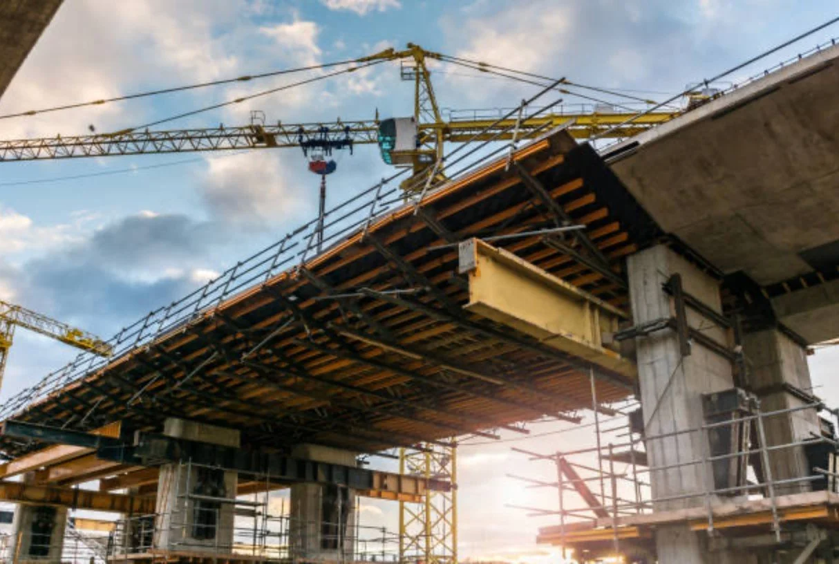 Construction site with a bridge under construction, scaffolding, and a yellow tower crane against a partly cloudy sky.