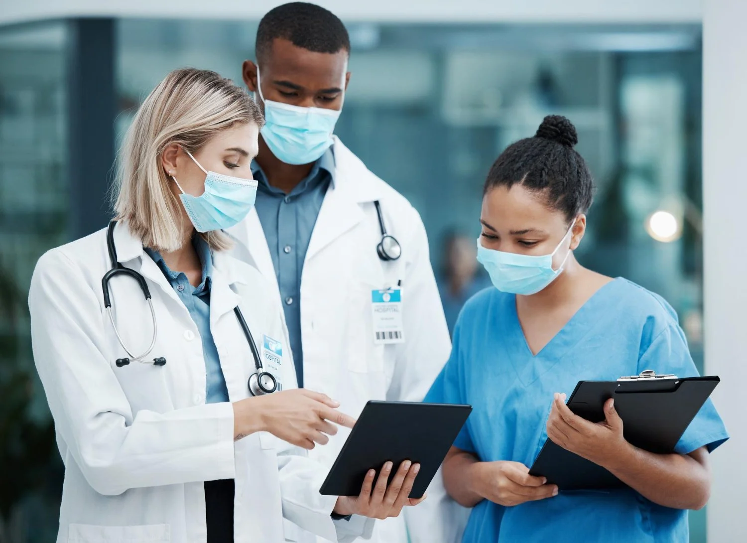 Three healthcare professionals, two women and one man, wearing face masks and medical scrubs, looking at tablets in a hospital lobby.