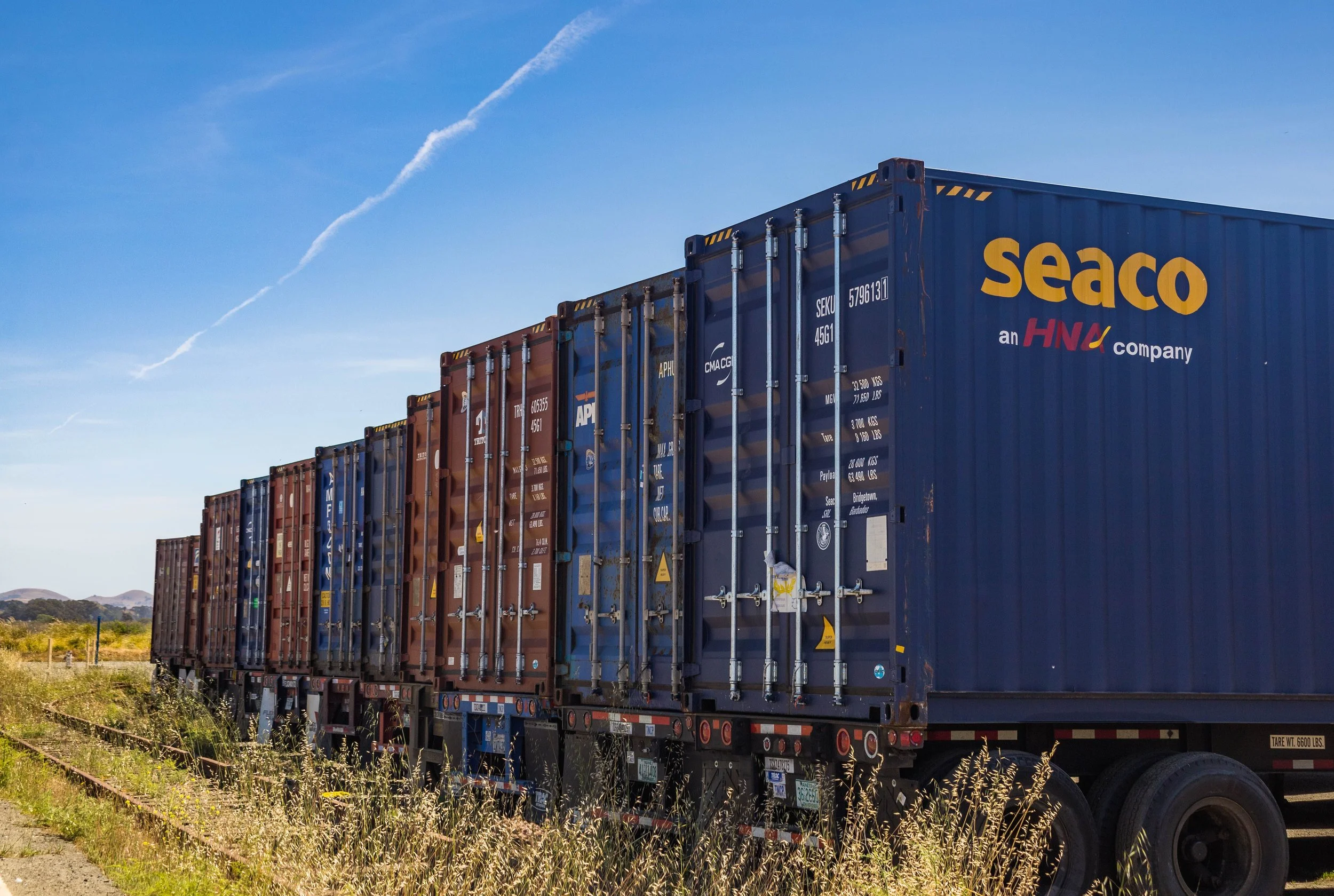 A line of colorful shipping containers on a flatbed truck traveling on a highway under a blue sky with a few clouds.