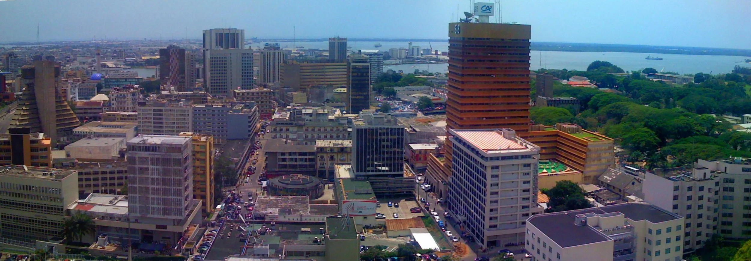 Aerial view of a cityscape showing numerous tall buildings, a river in the background, and areas of green trees, with some ships visible on the water.