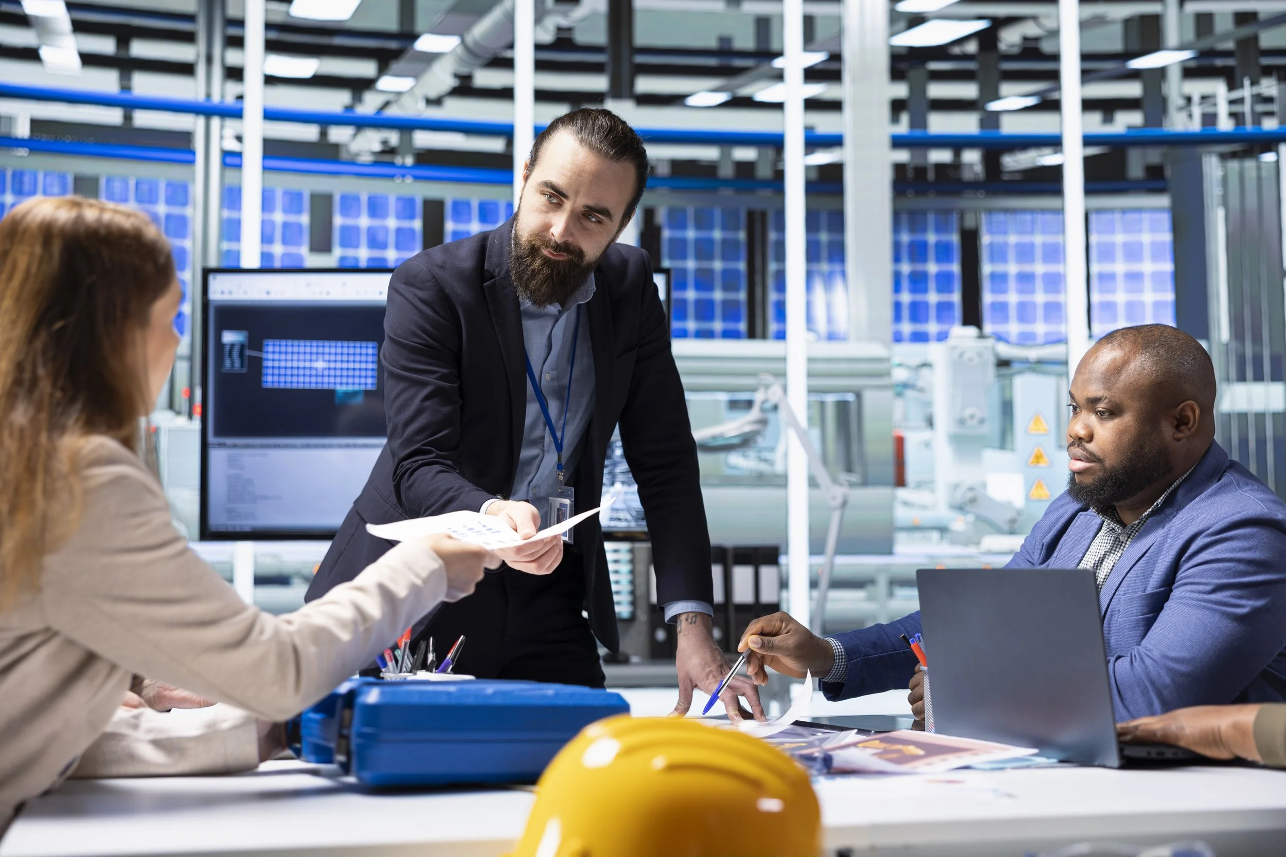 A group of four people in a modern technological workspace. One man with a beard and bun is leaning over a table, handing papers to a woman with long hair, while a man with a shaved head and a man with short hair and beard are seated, one with a laptop. The background features large screens and solar panel installations.