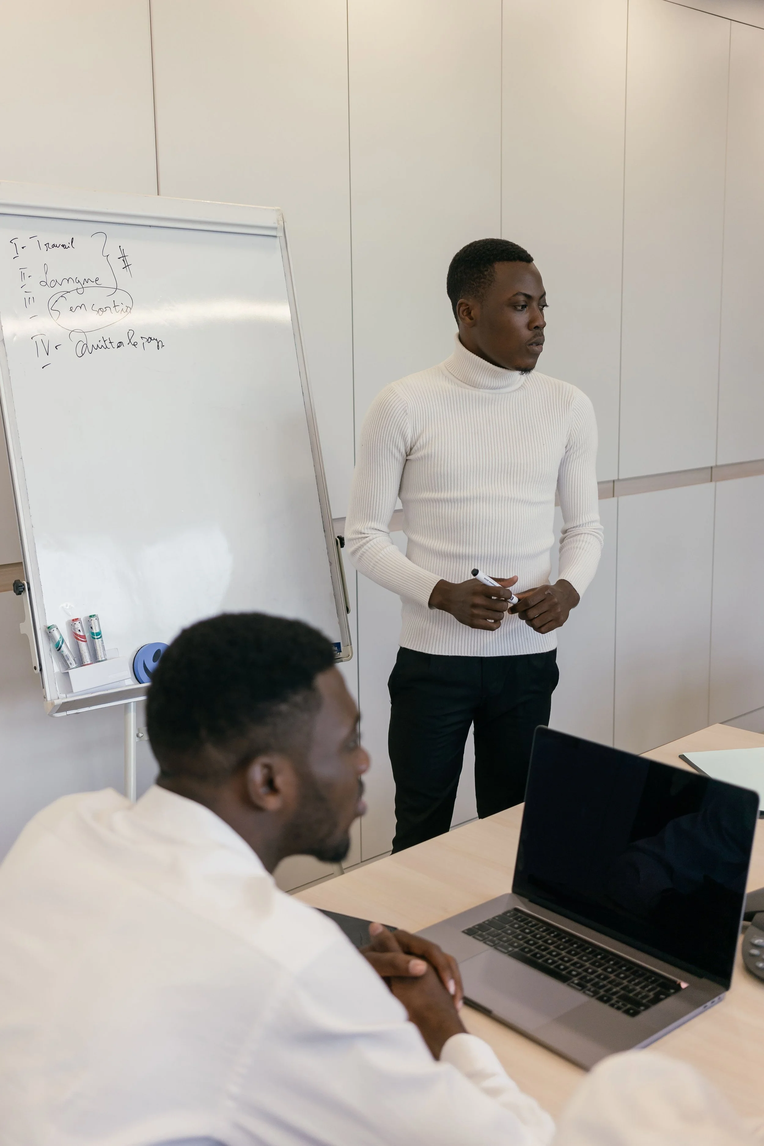 A man in a white turtleneck sweater standing in front of a whiteboard, holding a marker, and looking to the side during a presentation or class. Two seated men are at a table with laptops, one visible in the foreground and the other partially shown, both facing the presenter.