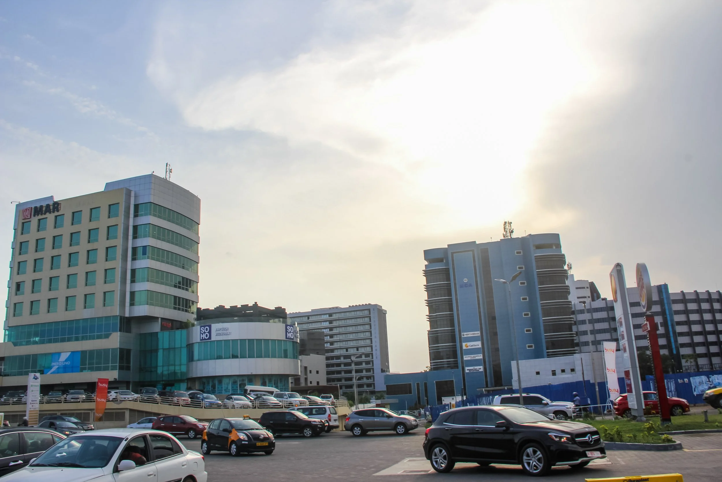 Cityscape featuring modern high-rise buildings, a parking lot with numerous cars, streetlights, and a partly cloudy sky with the sun partially obscured by clouds.