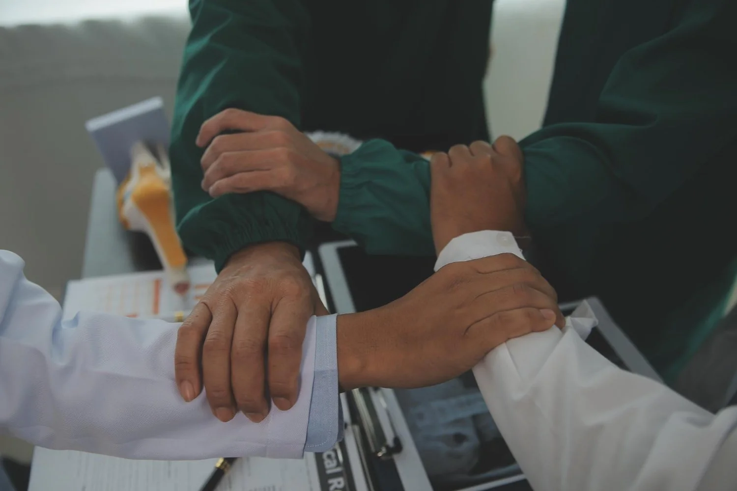 Multiple hands gripping each other's forearms in a gesture of support or teamwork, with a table of documents and electronic devices in the background.