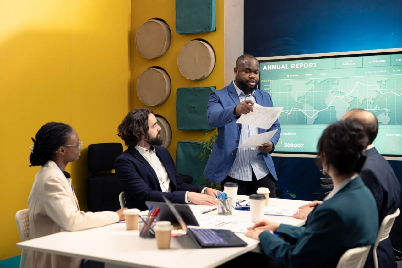 Business meeting in a conference room with a man standing and presenting a report on a large screen, while five seated colleagues watch.