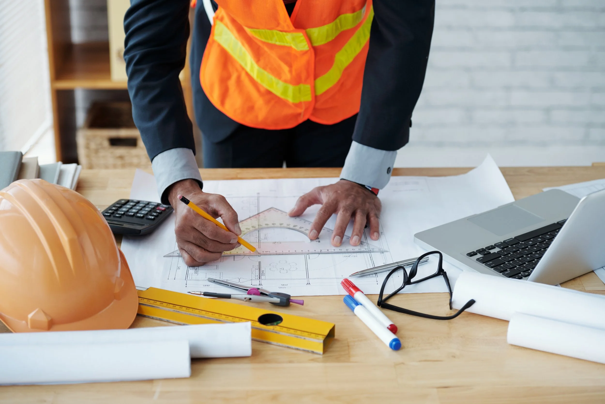 Person in safety vest and suit working on architectural plans at a desk, surrounded by a calculator, pens, glasses, a yellow level, a hard hat, and a laptop.