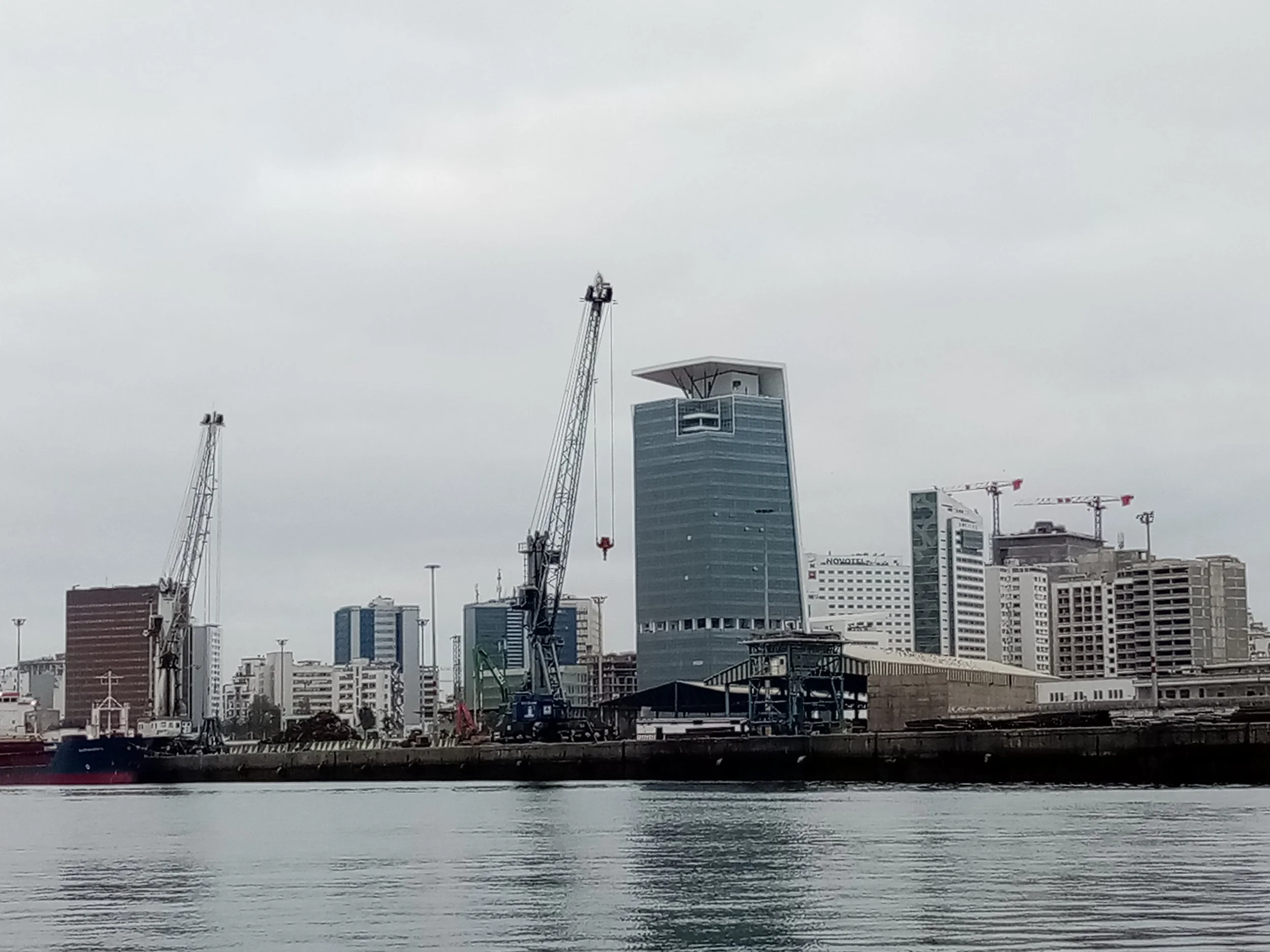 Cityscape of tall modern buildings under cloudy sky, view from across the water with construction cranes visible.