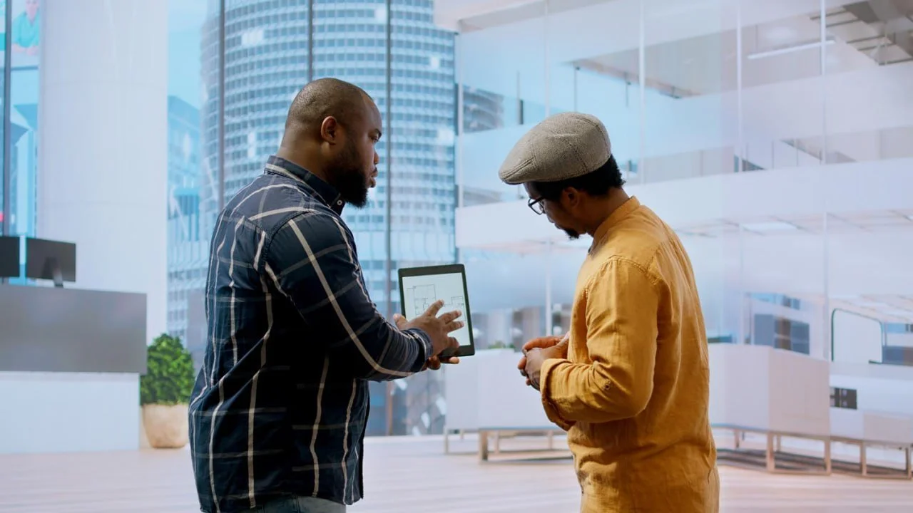 Two men standing indoors with a cityscape visible through large windows; one man is showing something on a tablet to the other.