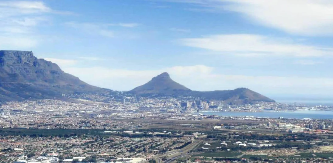 Panoramic view of Cape Town with Table Mountain and surrounding cityscape under a partly cloudy sky.