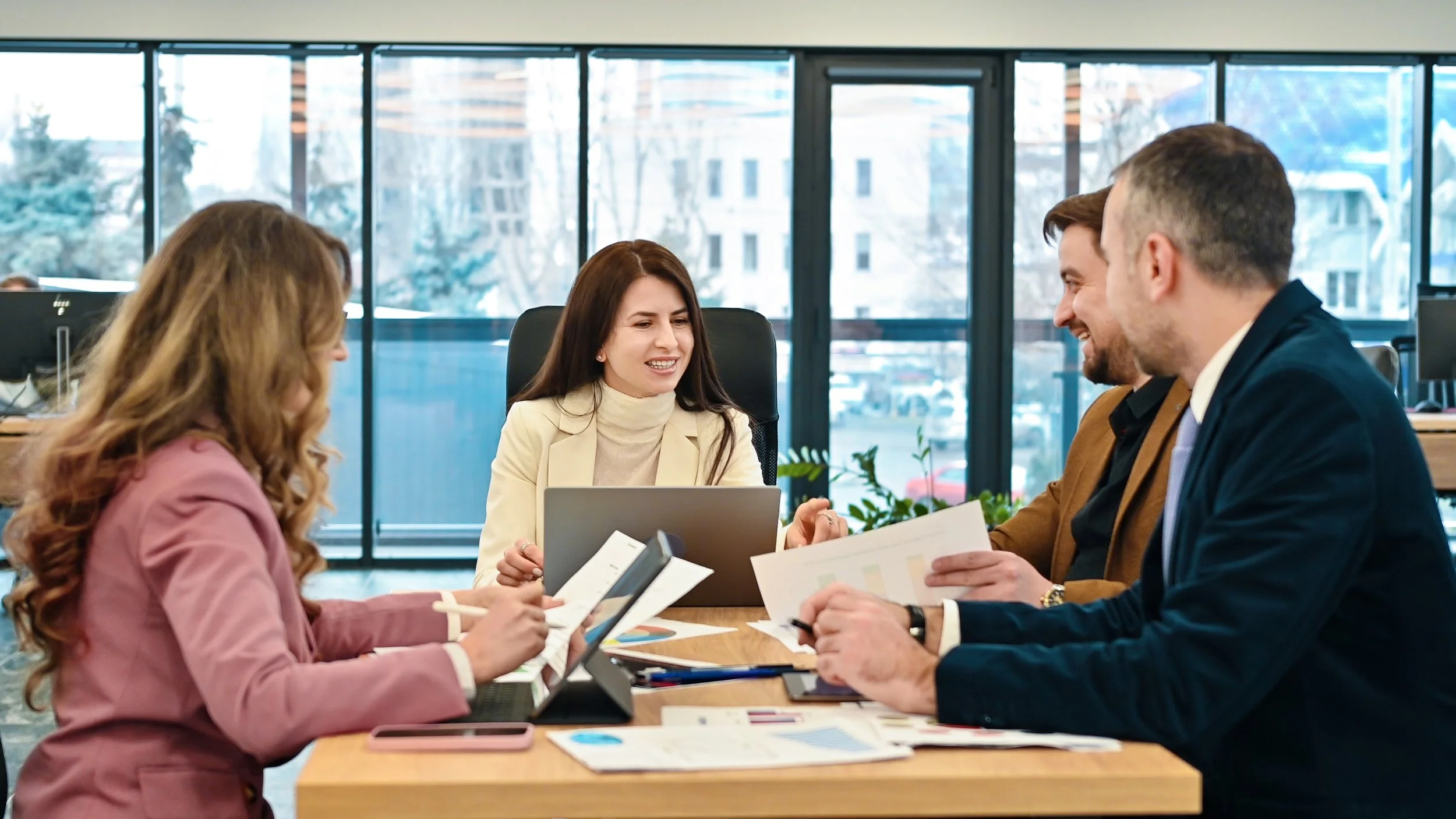 A group of four professionals in business attire having a meeting around a table in an office with large windows showing an urban landscape outside.