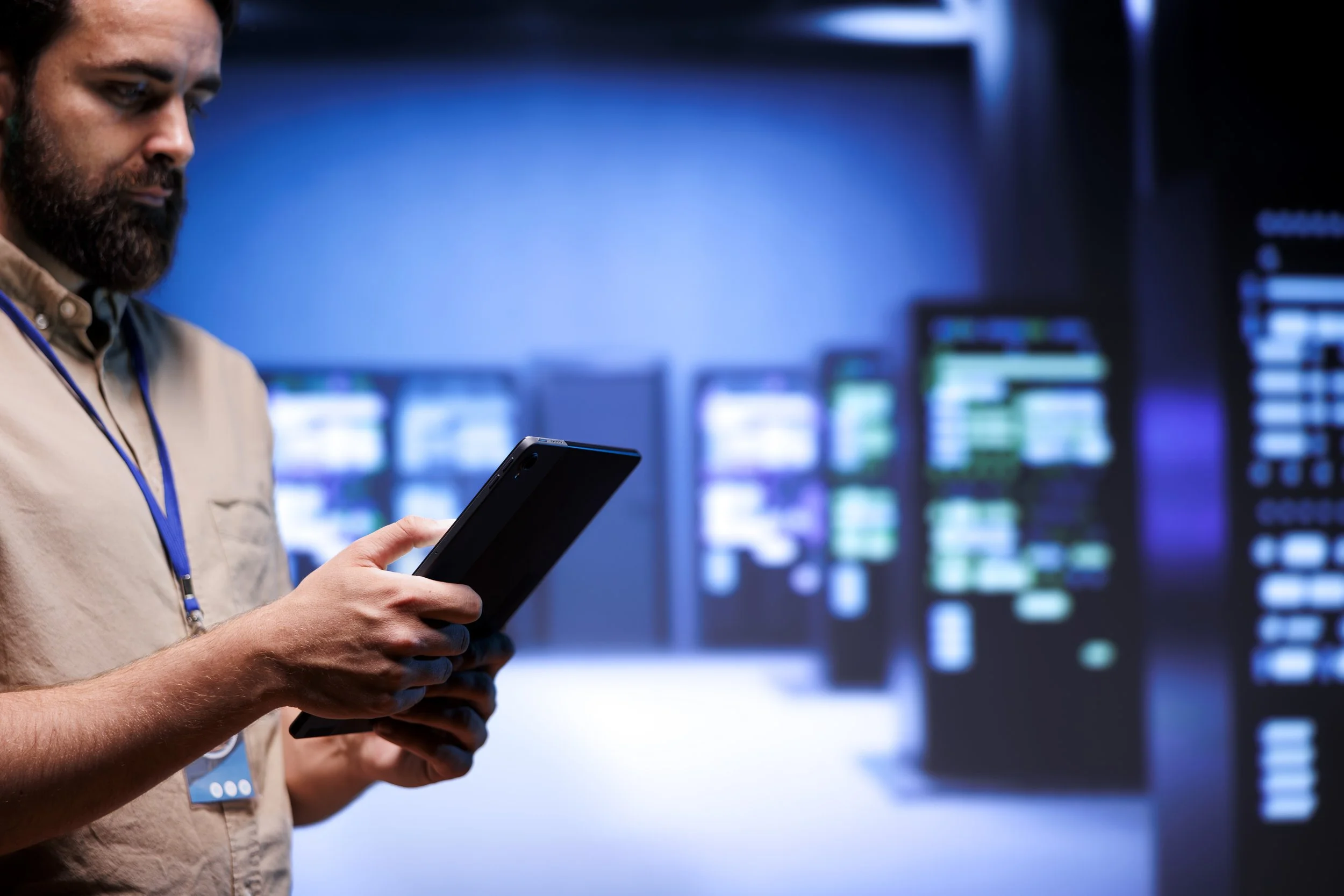 A man with a beard wearing a beige shirt and a lanyard stands in front of multiple computer screens, holding and looking at a tablet in his hands in a dimly lit room with blue lighting.