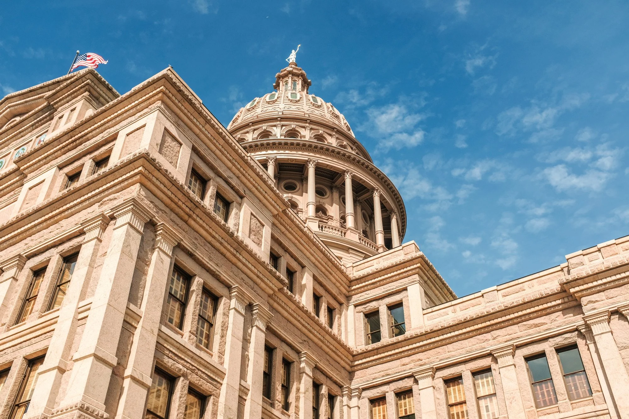 Close-up of a historic government building with a large dome topped with a statue, set against a bright blue sky with wispy clouds.