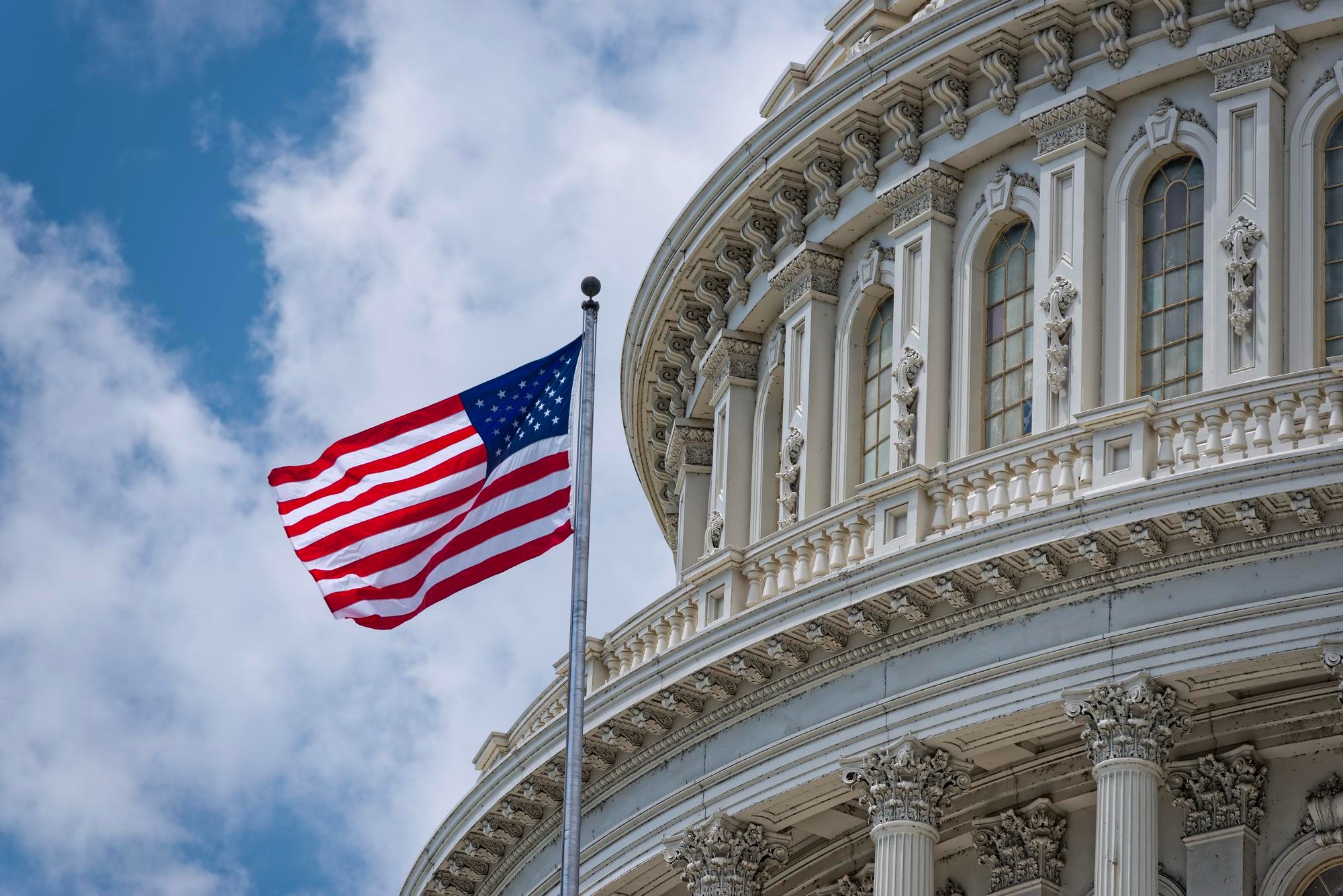 The United States Capitol building with an American flag flying in front, showing detailed neoclassical architecture with columns and decorative elements, against a partly cloudy sky.