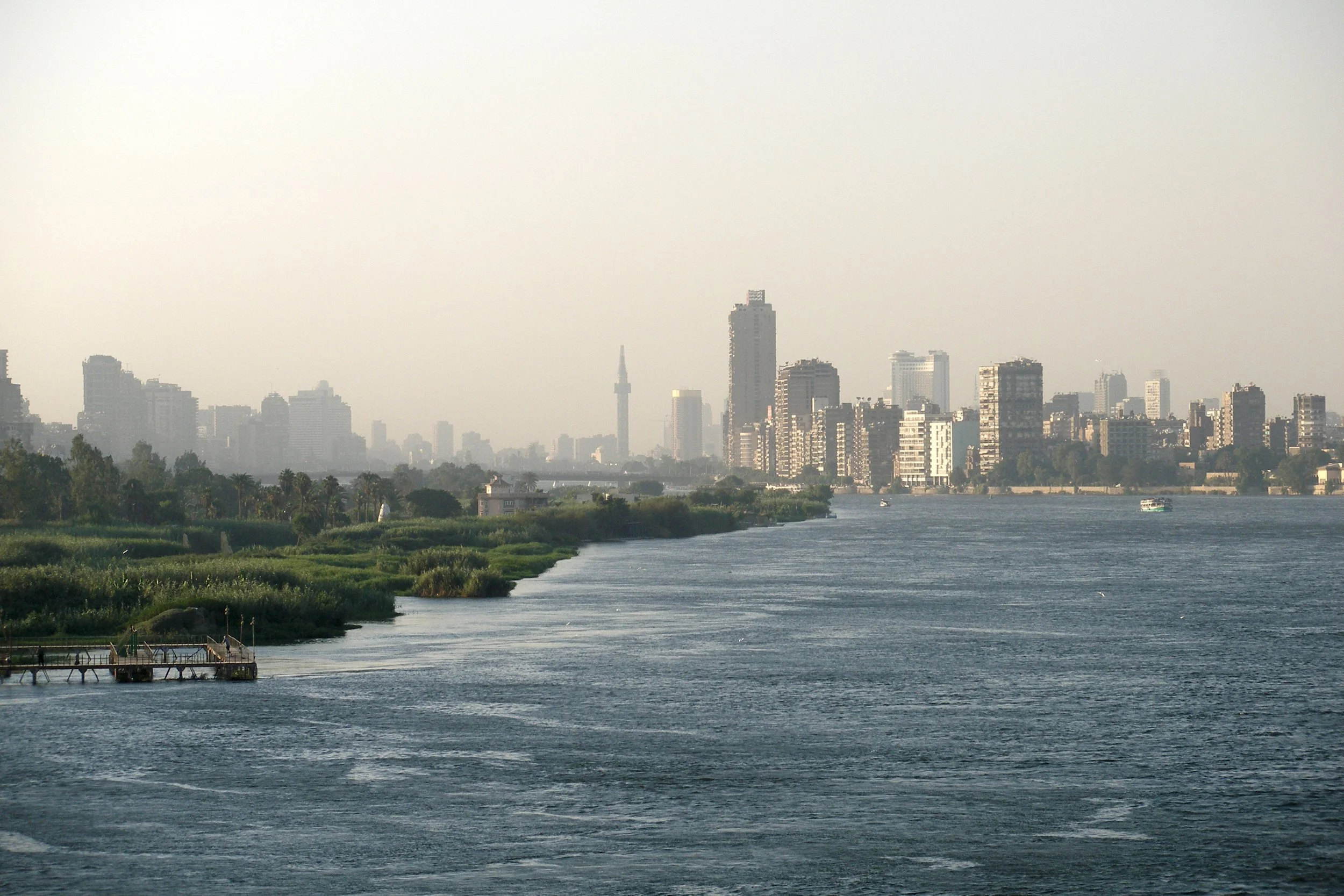 A city skyline with tall buildings in the background and a river in the foreground, with a small dock extending into the water.