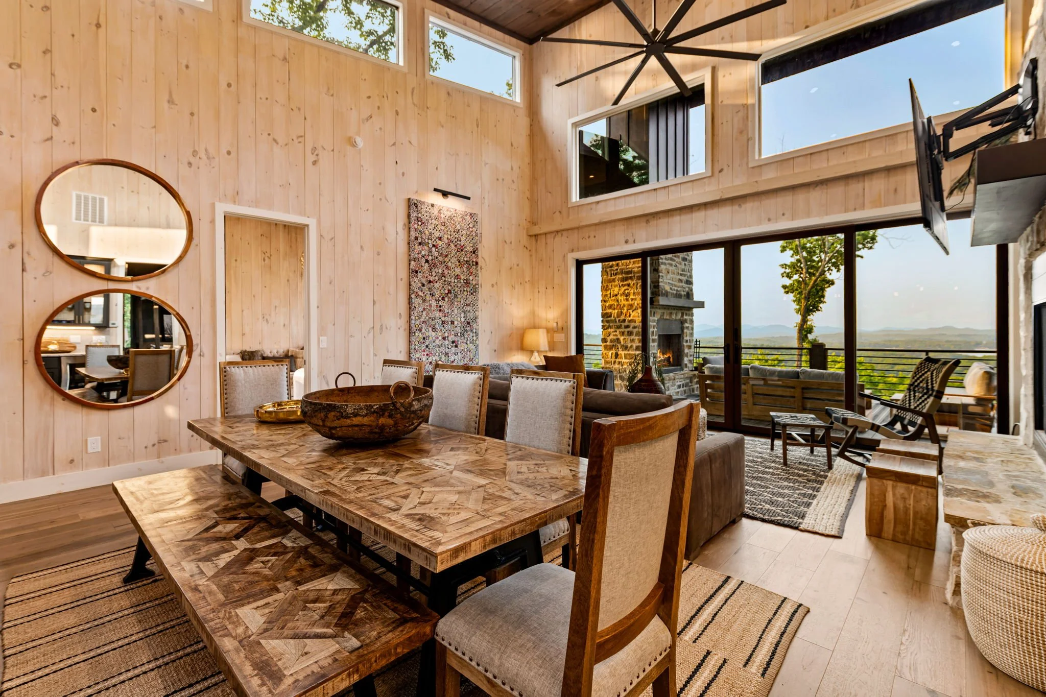 Gather in this elegant dining space featuring a large farmhouse table, bench seating, designer lighting, vaulted ceilings, Smart TV, and floor-to-ceiling glass doors opening to a scenic deck with breathtaking mountain and lake views.