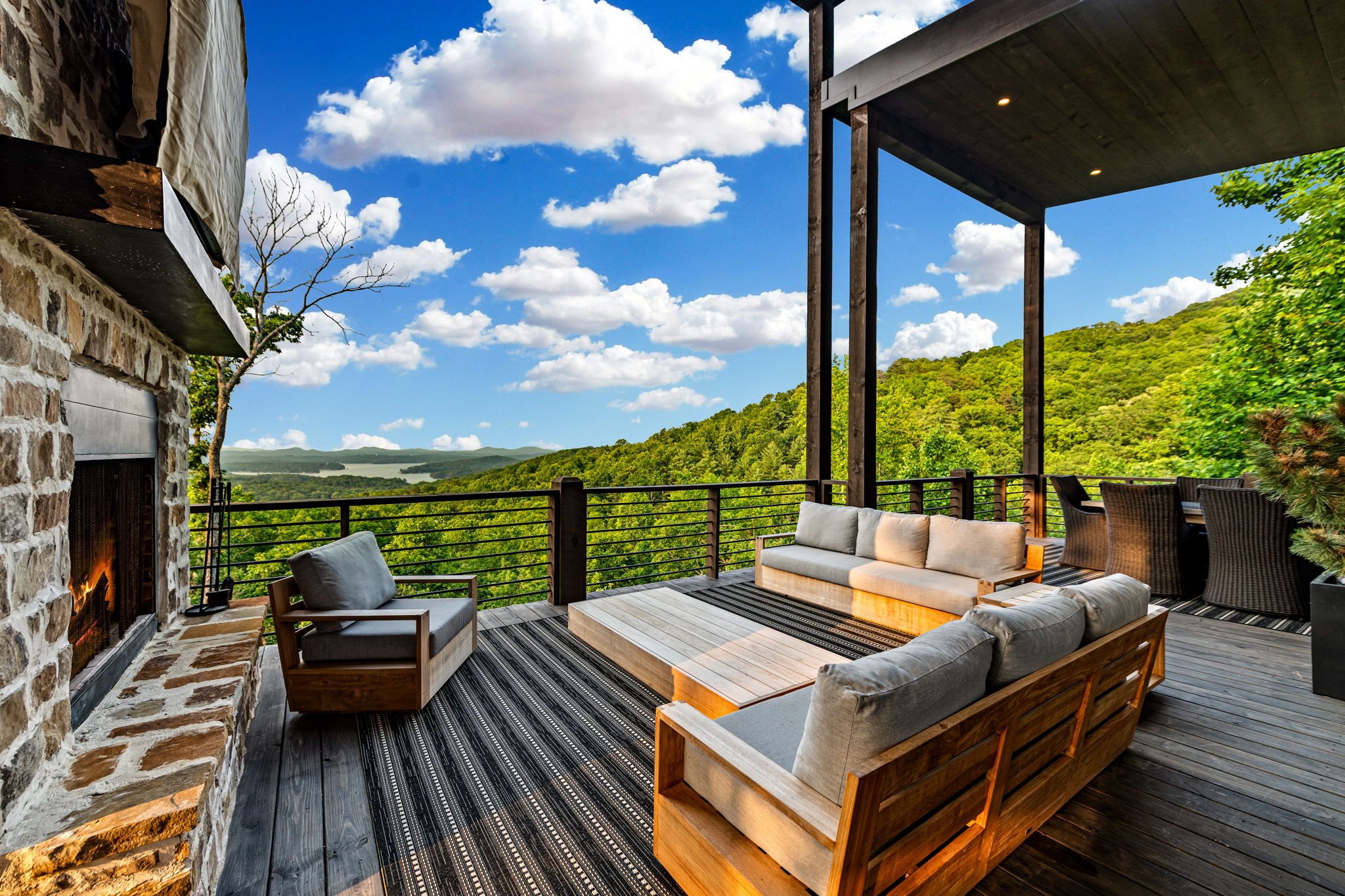 Unwind on the expansive covered deck featuring outdoor lounge seating, a stone fireplace, and panoramic mountain & lake views—a luxurious open-air space perfect for relaxing, entertaining, and enjoying peaceful sunsets.
