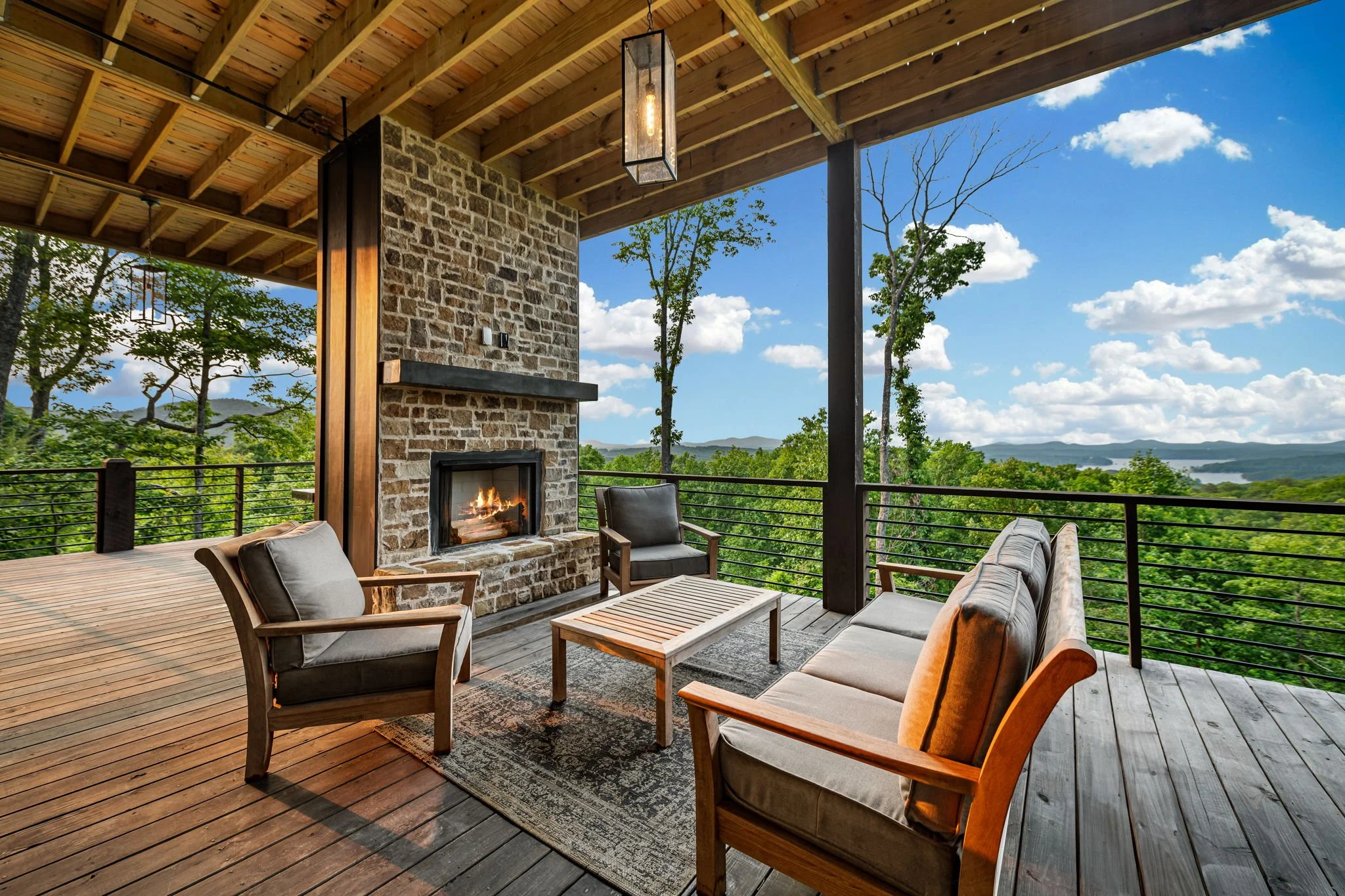 Relax on the covered deck featuring a cozy stone fireplace, outdoor lounge seating, and breathtaking panoramic mountain & lake views—the perfect luxury setting for morning coffee, sunset cocktails, or peaceful evenings in the fresh mountain air.