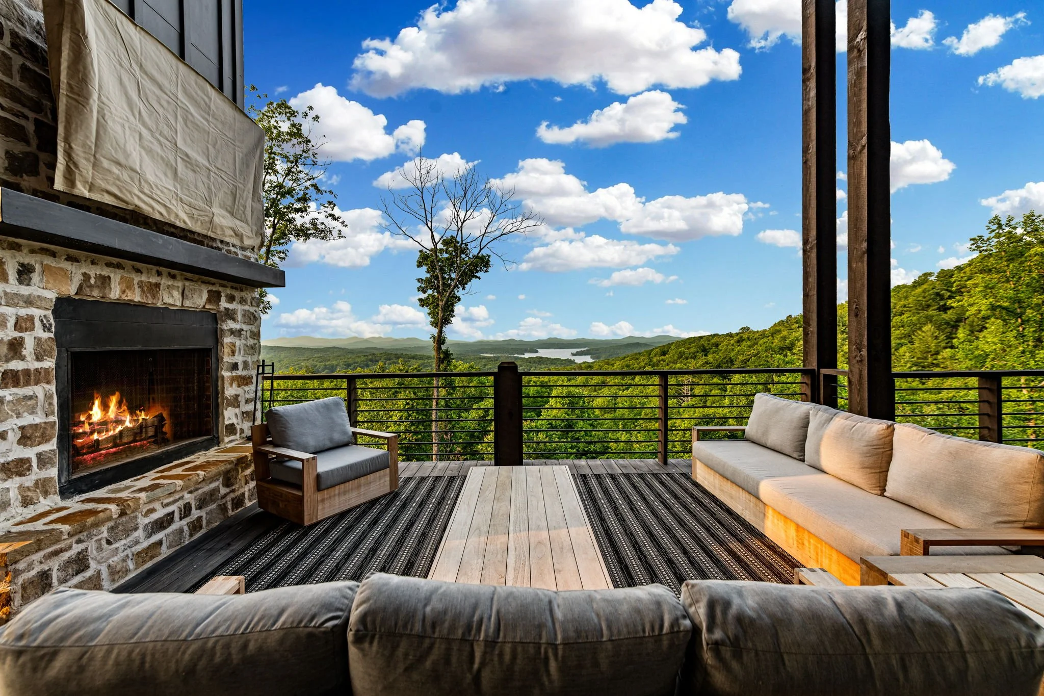 Relax by the outdoor stone fireplace on this spacious deck featuring comfortable lounge seating and sweeping mountain & lake views—a serene luxury setting perfect for cozy evenings and unforgettable sunsets.