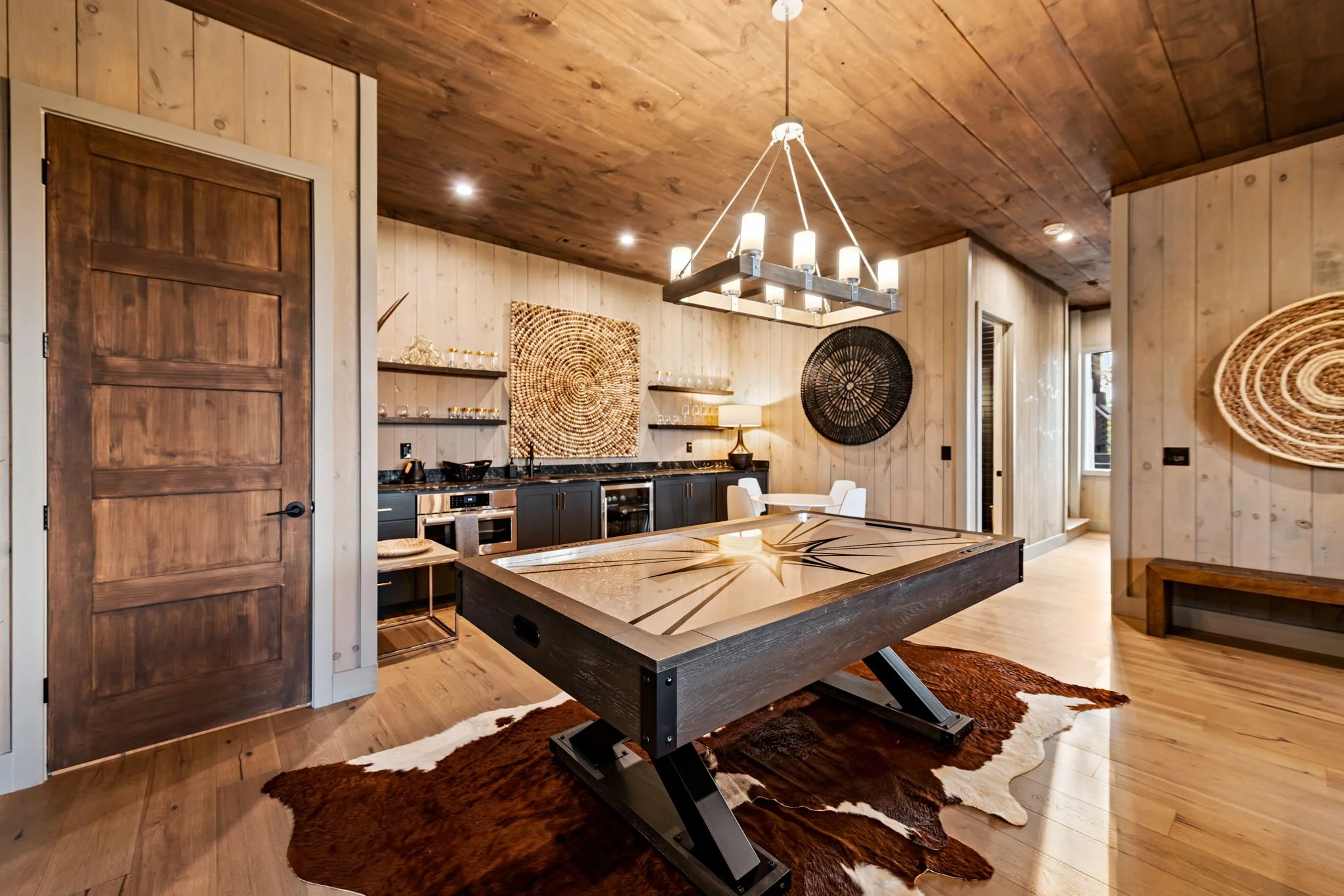 Play & unwind in this stylish game area featuring a premium shuffleboard table, wet bar with sink, beverage fridge, open shelving, designer lighting, and lounge seating—a perfect space for entertaining, games, and evening cocktails.