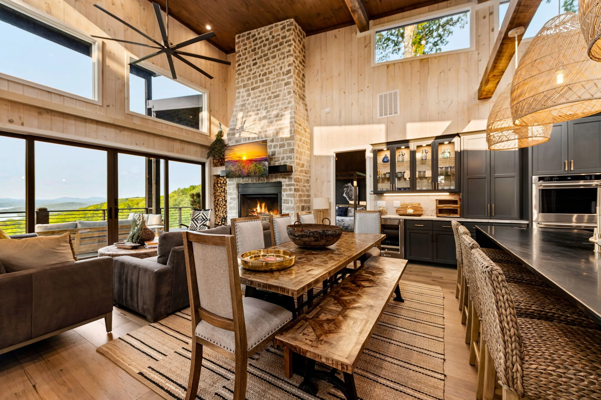 Gather in this stunning open-concept space featuring a large dining table with bench seating, chef’s kitchen with island seating, custom cabinetry, designer pendant lighting, vaulted ceilings, and a floor-to-ceiling stone fireplace w/ mountain views.