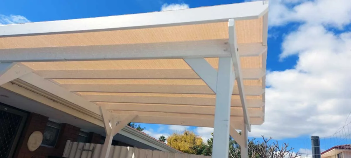 White pergola with beige shading panels over a backyard patio, with a blue sky and clouds in the background.