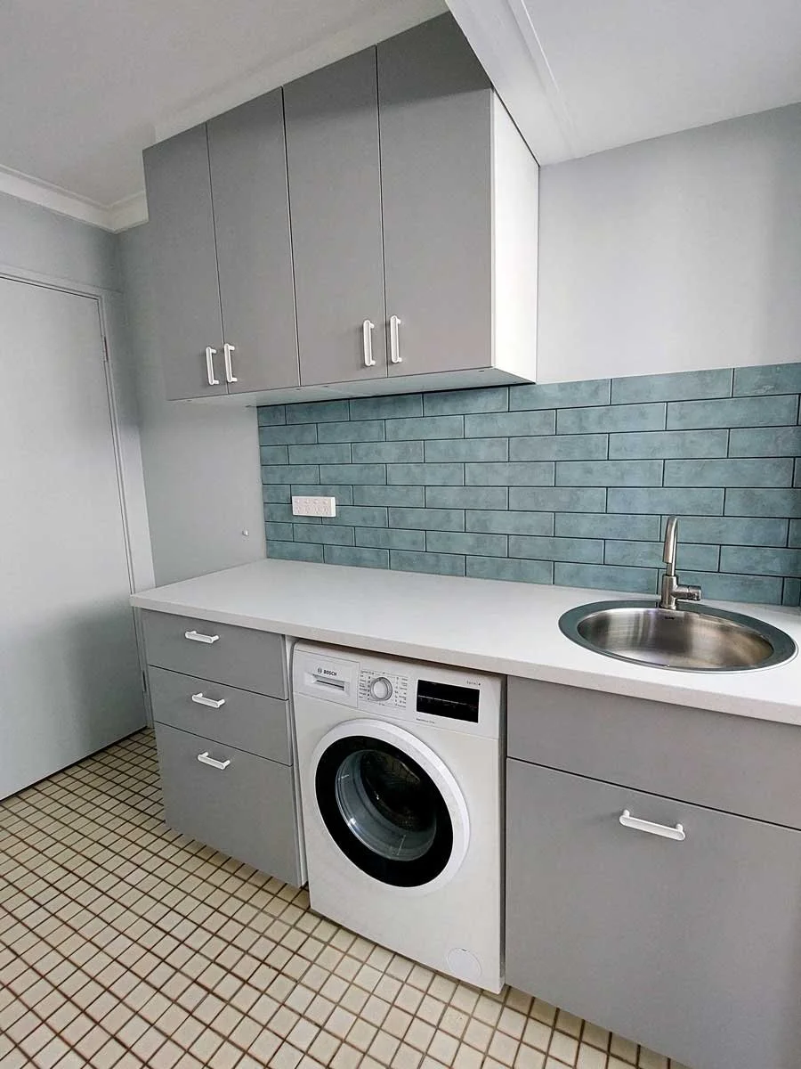 A Laundry with gray Ikea cabinets, a white countertop, a small stainless steel sink, and a washing machine beneath the counter. There are blue brick-style tiles on the wall behind the counter and a tiled floor.