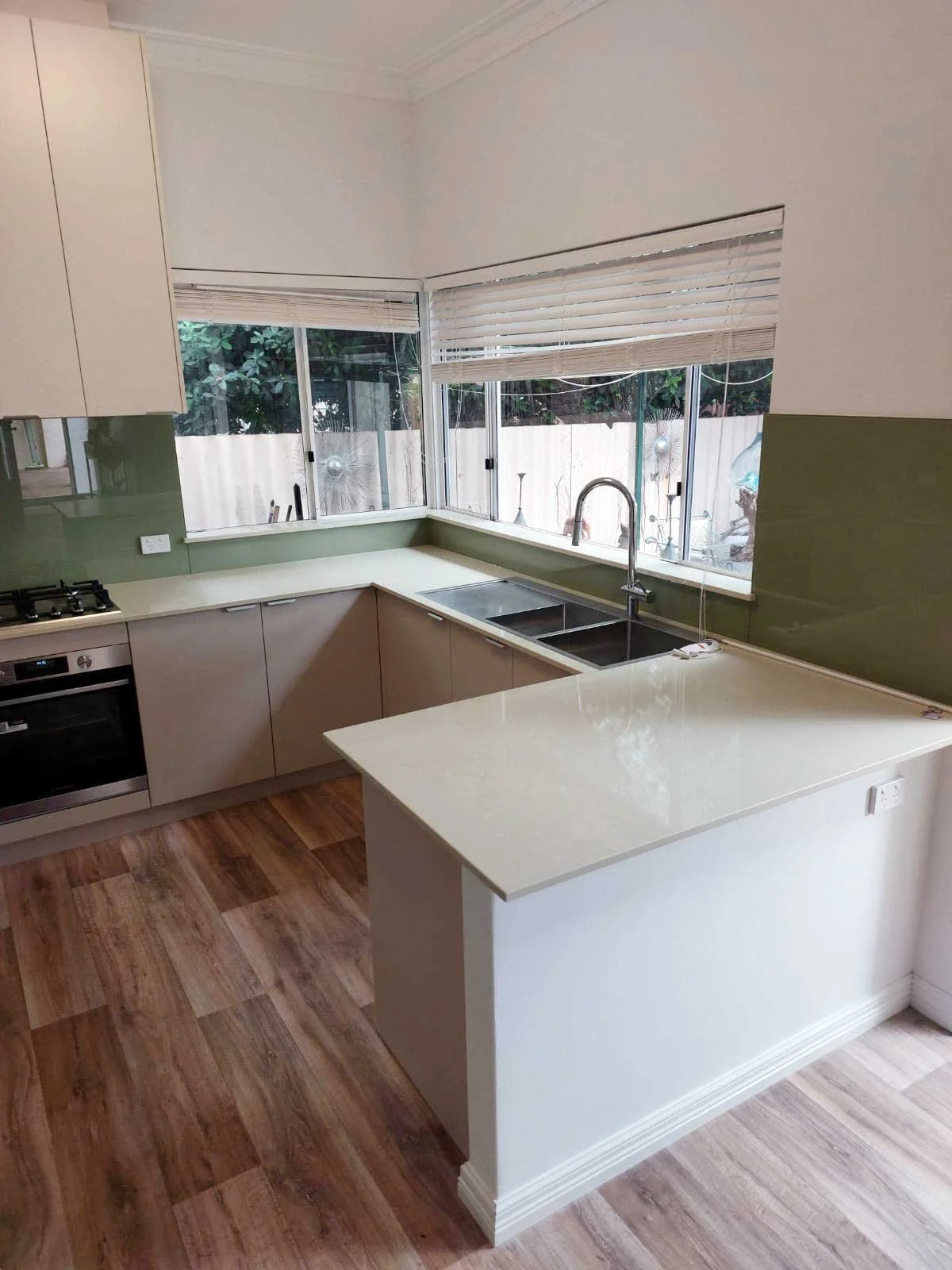 Modern kitchen with white cabinets, a green backsplash, and a large window with blinds, featuring a double sink and a built-in oven.
