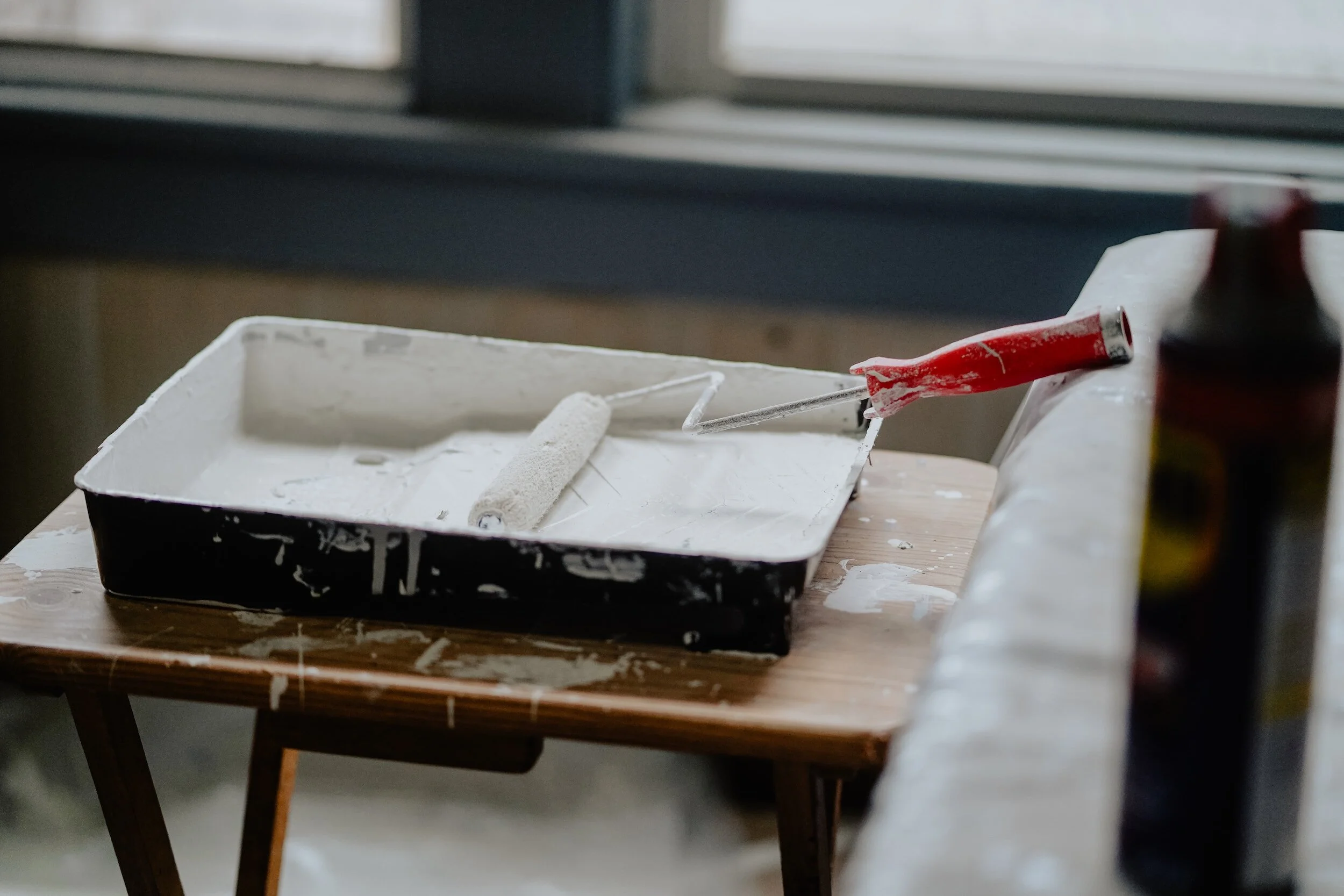 Paint roller, brushes, and a paint tray on a wooden table, with paint on the roller and tray in a room near a window.
