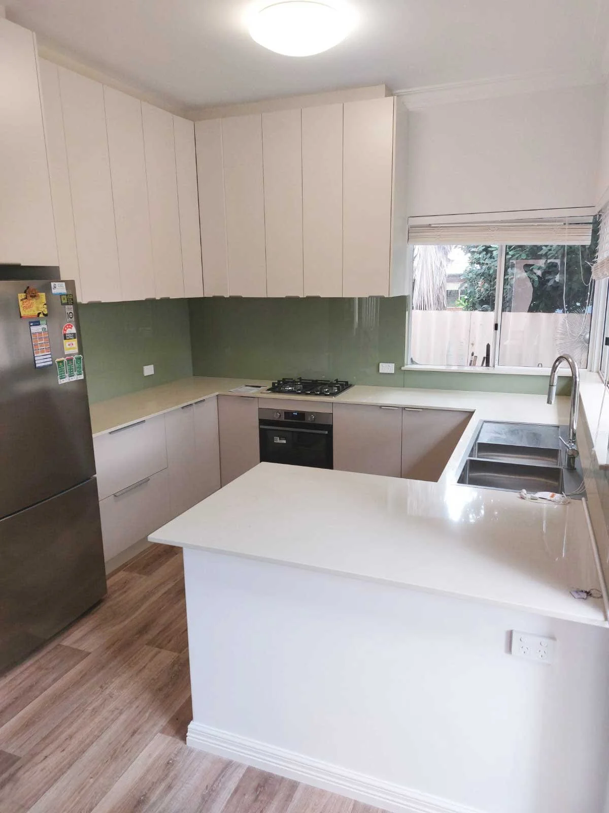Modern kitchen with white cabinets, green backsplash, stainless steel refrigerator, built-in oven, gas stove, and double sink. Bright natural light from window with view of trees.