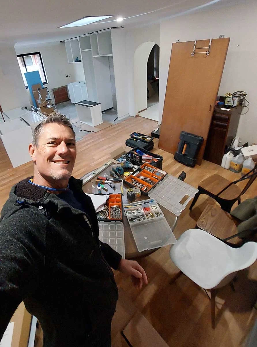 Paul smiling at a camera during a kitchen renovation, with various tools and supplies on a dining table, in a room with hardwood floors, partially installed white Ikea cabinets, and a doorway leading to a laundry area.