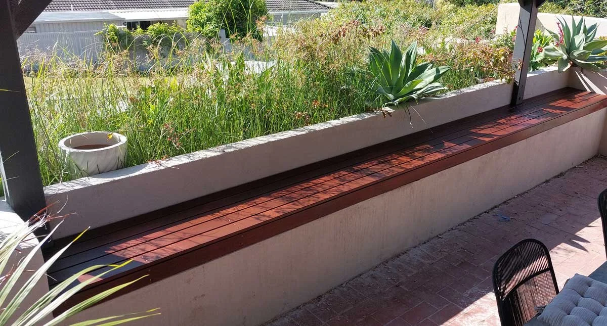 A balcony garden with various green plants, including succulents and grasses, in a white planter box. There is brick flooring and chairs are visible in the foreground.