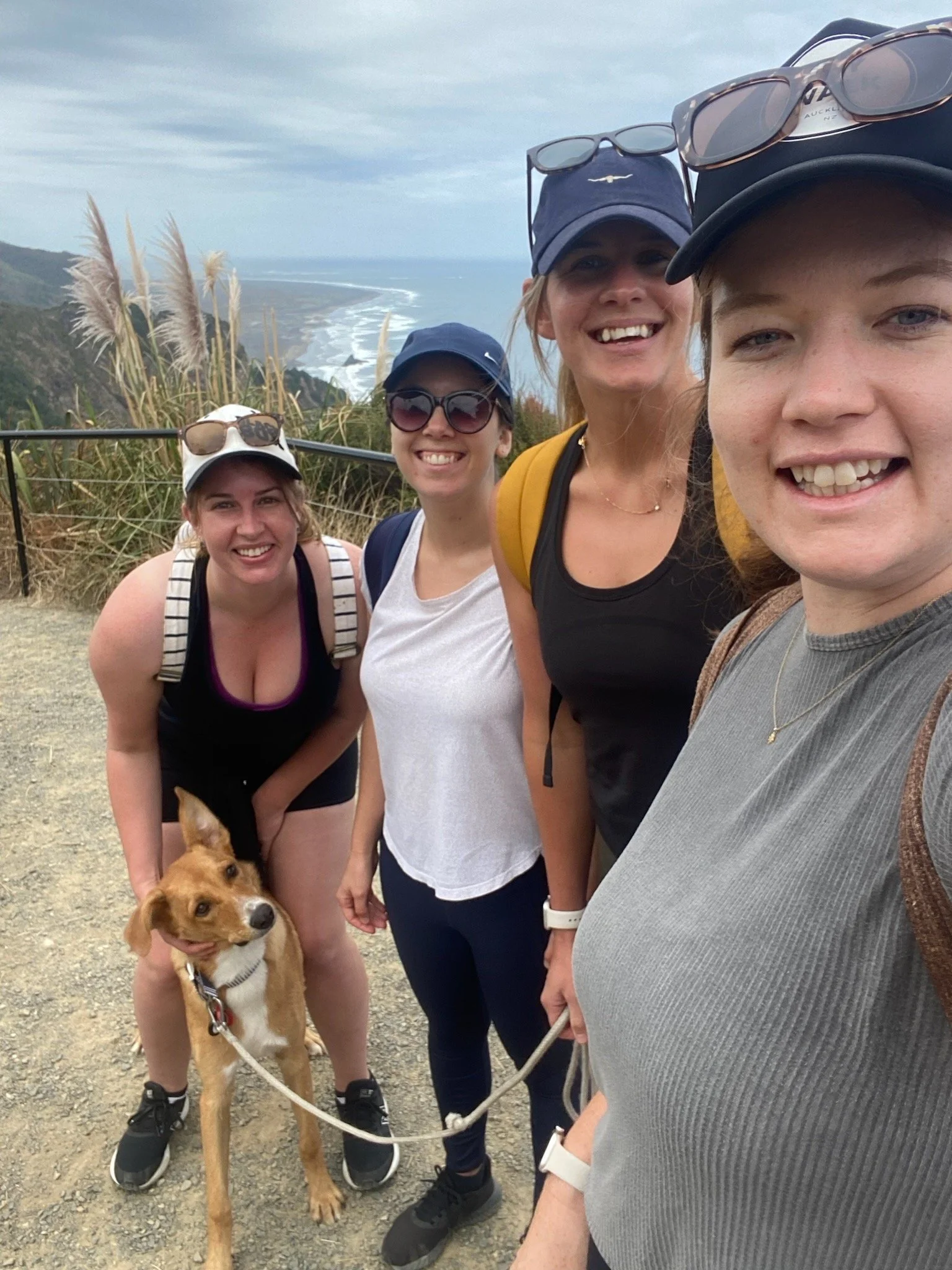 Amy Callon with Friends with sunglasses and hats smiling for a selfie with a dog at the beach. The background shows coastal cliffs, ocean waves, and cloudy sky.