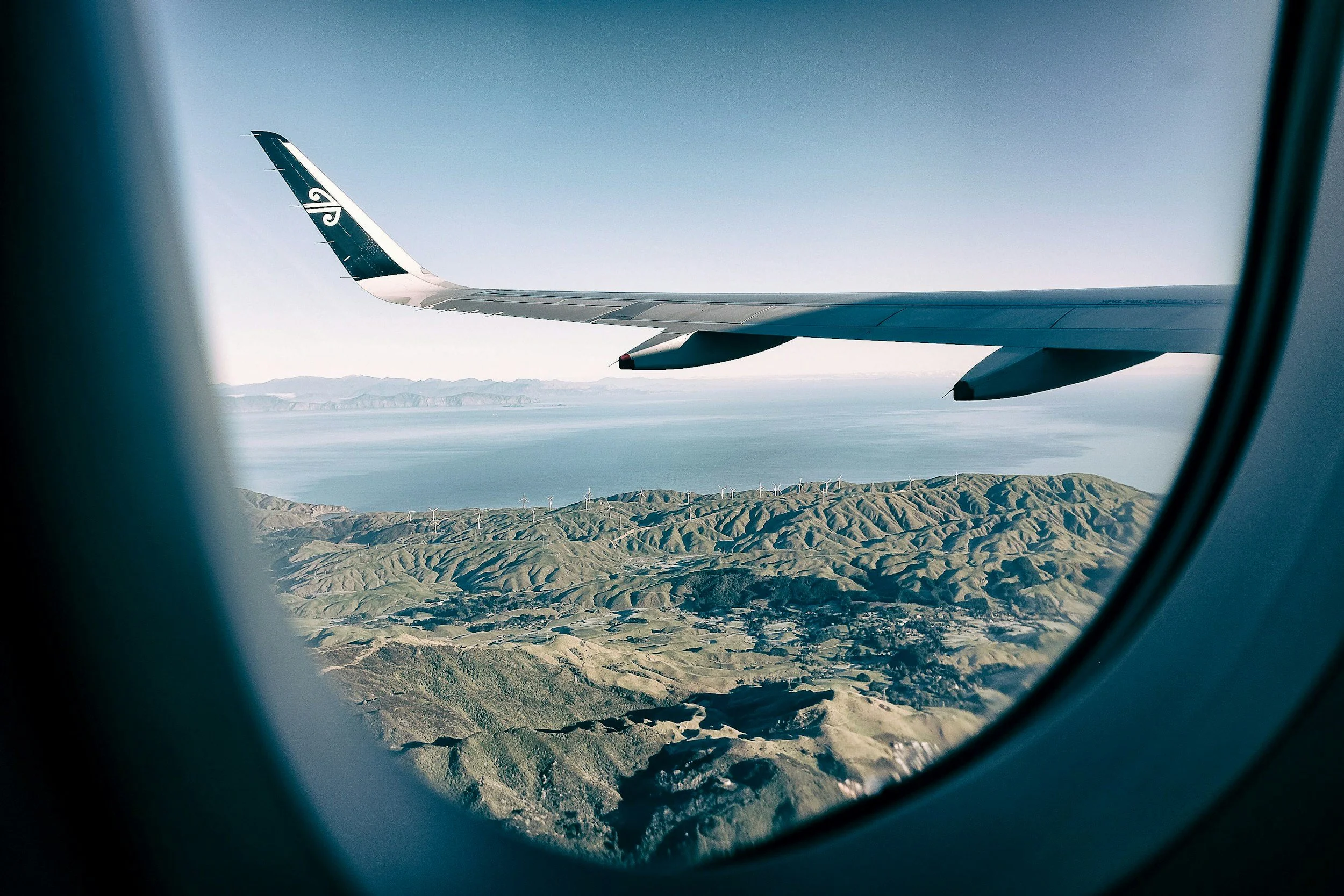 View from an airplane window showing airplane wing, mountainous landscape, wind turbines, and ocean in the background.