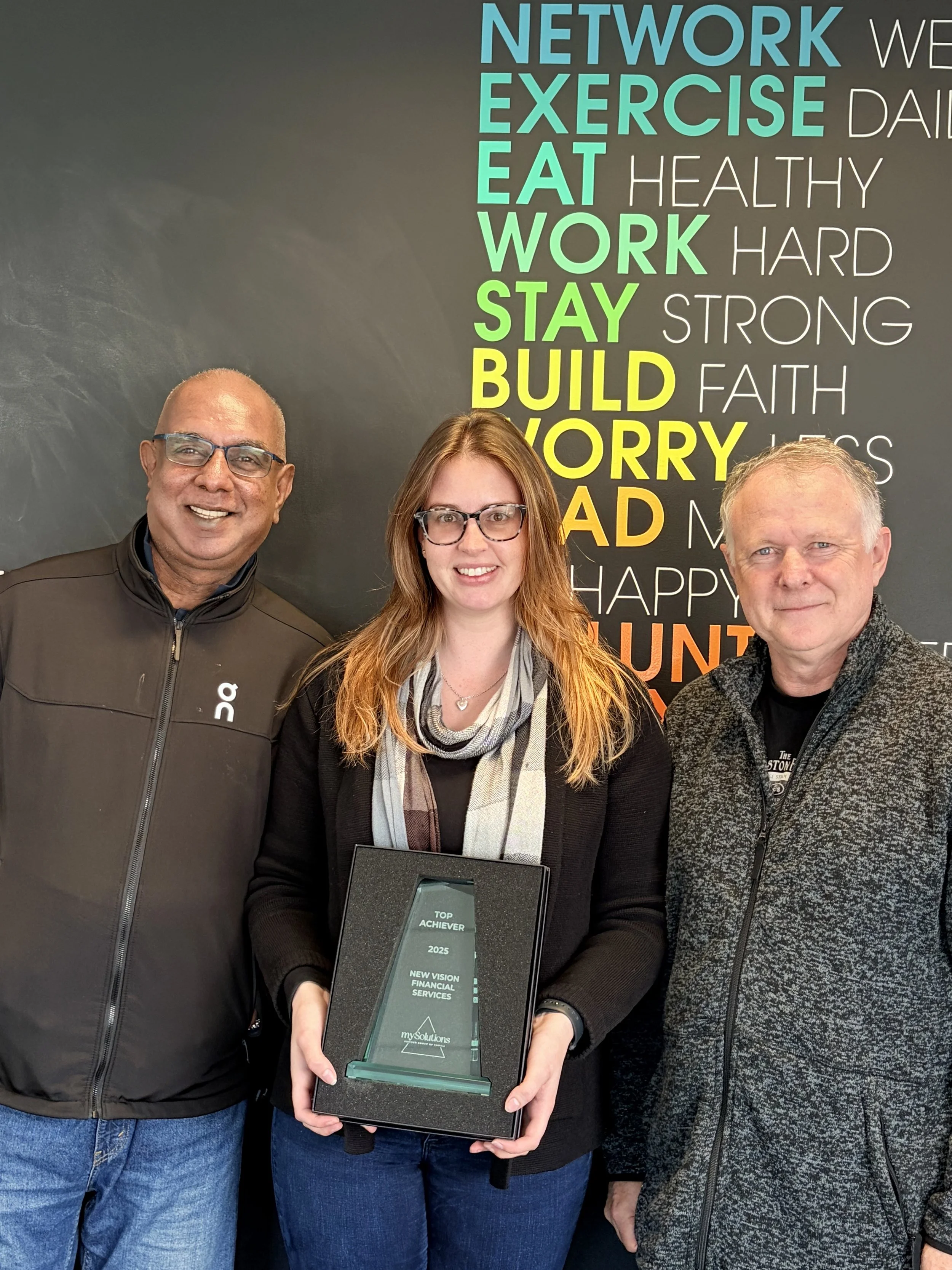 New Vision Team, Nimalka Perera, Amy Callon and Glen Hatcher, in front of a black wall. Amy holding a glass award that reads 'TOP ACHIEVER 2025' for 'NEW VISION FINANCIAL SERVICES.'
