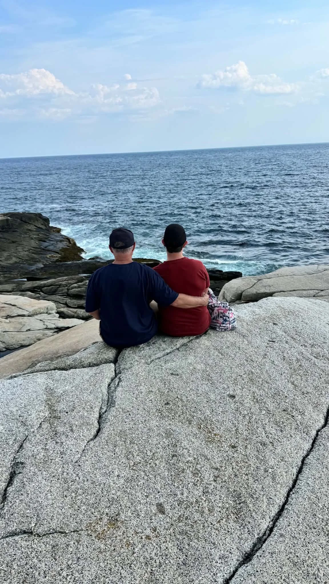 Two people sitting on rocks by the ocean, embracing and looking out at the water under a blue sky with scattered clouds.