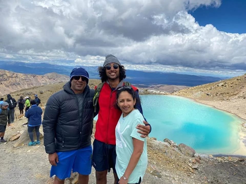 Nimalka Perera posing together outdoors near a turquoise crater lake with mountainous terrain and cloudy sky in the background.