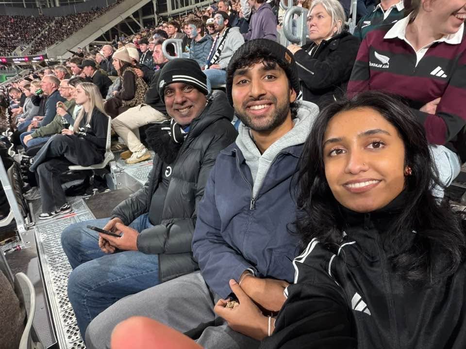 Nimalka Perera with Family sitting in stadium seats at a sports event, smiling for the camera, with spectators in the background.