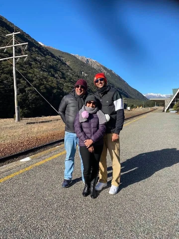 Nimalka Perera with Family standing outdoors near train tracks with mountains and snow-capped peaks in the background, smiling at the camera on a sunny day.
