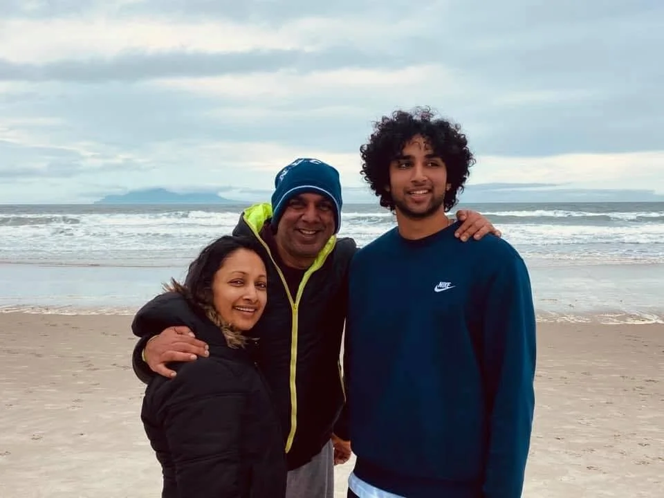 Nimalka Perera with Family smiling on a beach with waves in the background, dressed in jackets and hoodie.