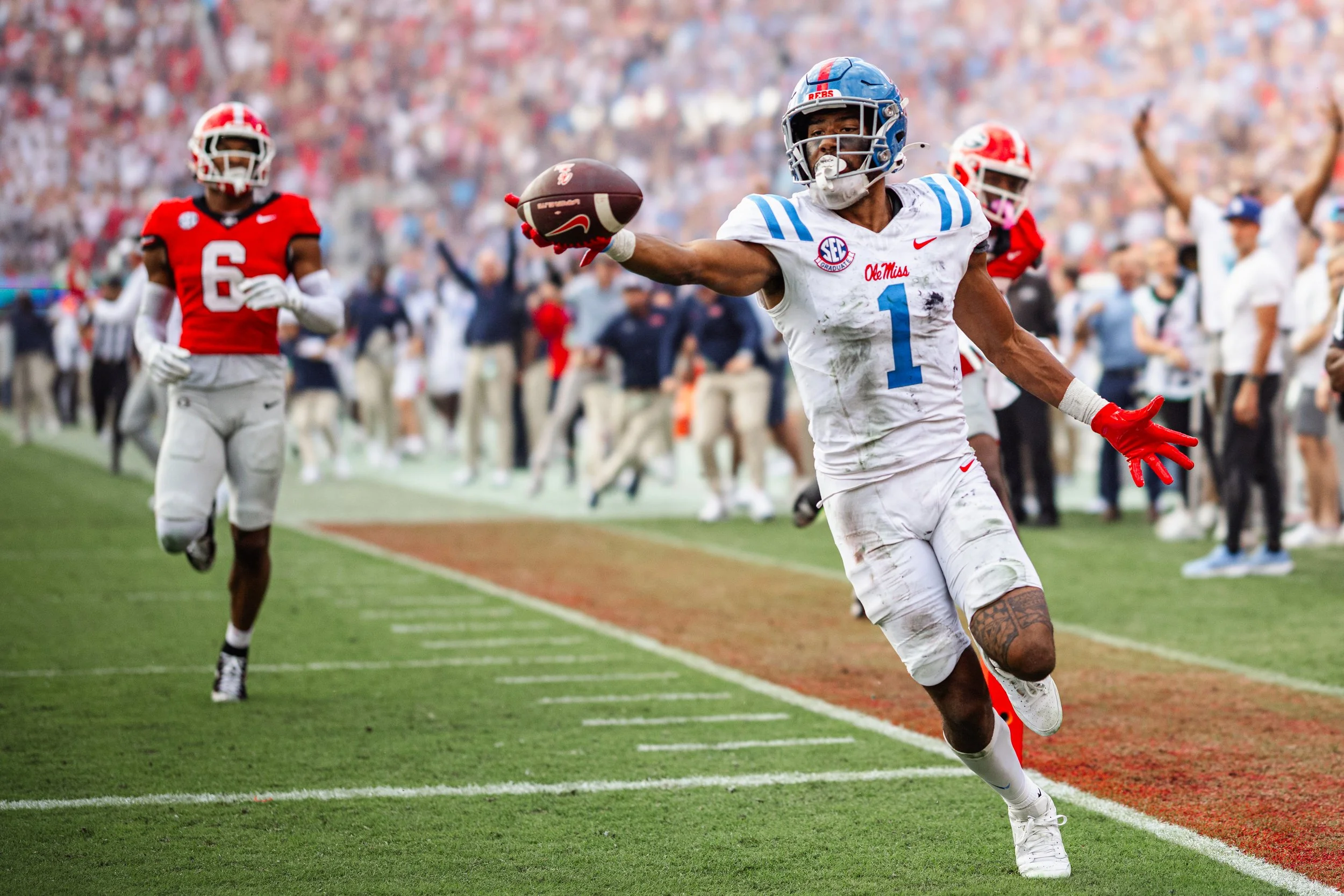Dezhuan striblinb of ole miss with the number 1 is running and catching a football in the end zone. Another player in a red uniform is approaching in the background, and there are spectators on the sidelines.