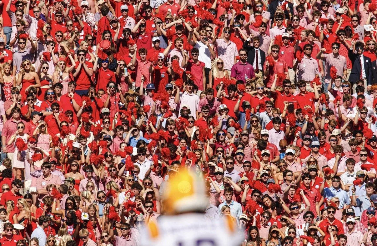 Crowd of sports fans dressed mostly in red, cheering and waving pom-poms at a stadium, with a blurred football in the foreground.
