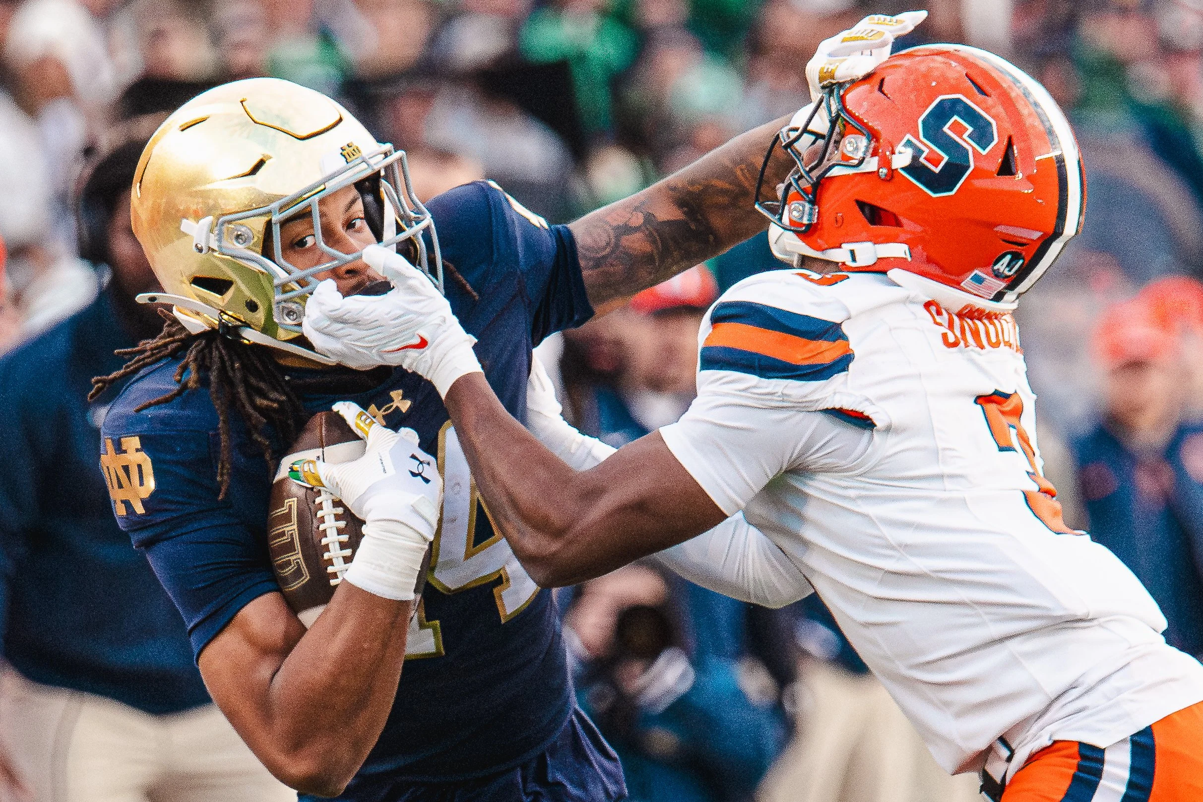 Two football players are engaged in a tackle during a game. The player on the left, wearing a navy blue and gold helmet and uniform, holds a football tightly while the player on the right, in an orange and white uniform with an orange helmet, tries t