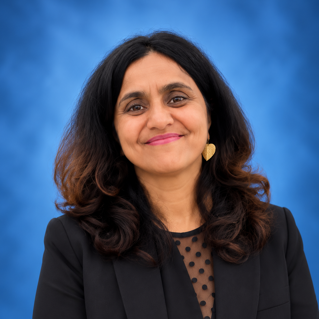 Portrait of a woman with dark, wavy hair wearing a black blazer and a sheer top with polka dots, gold leaf-shaped earrings, smiling against a blue background.
