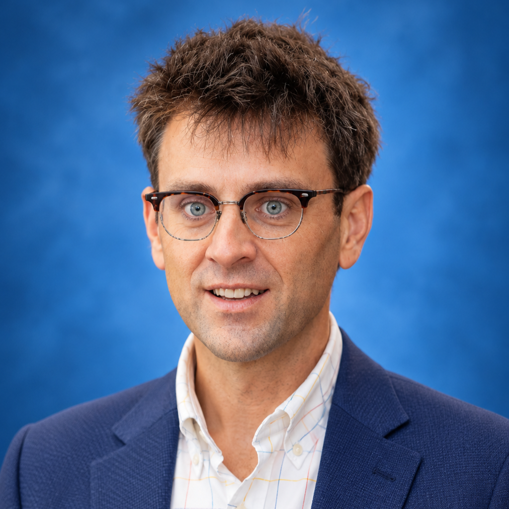 A man with light skin, blue eyes, and dark, slightly messy hair wearing glasses, a dark blue blazer, and a white shirt with a pattern, standing against a blue background.