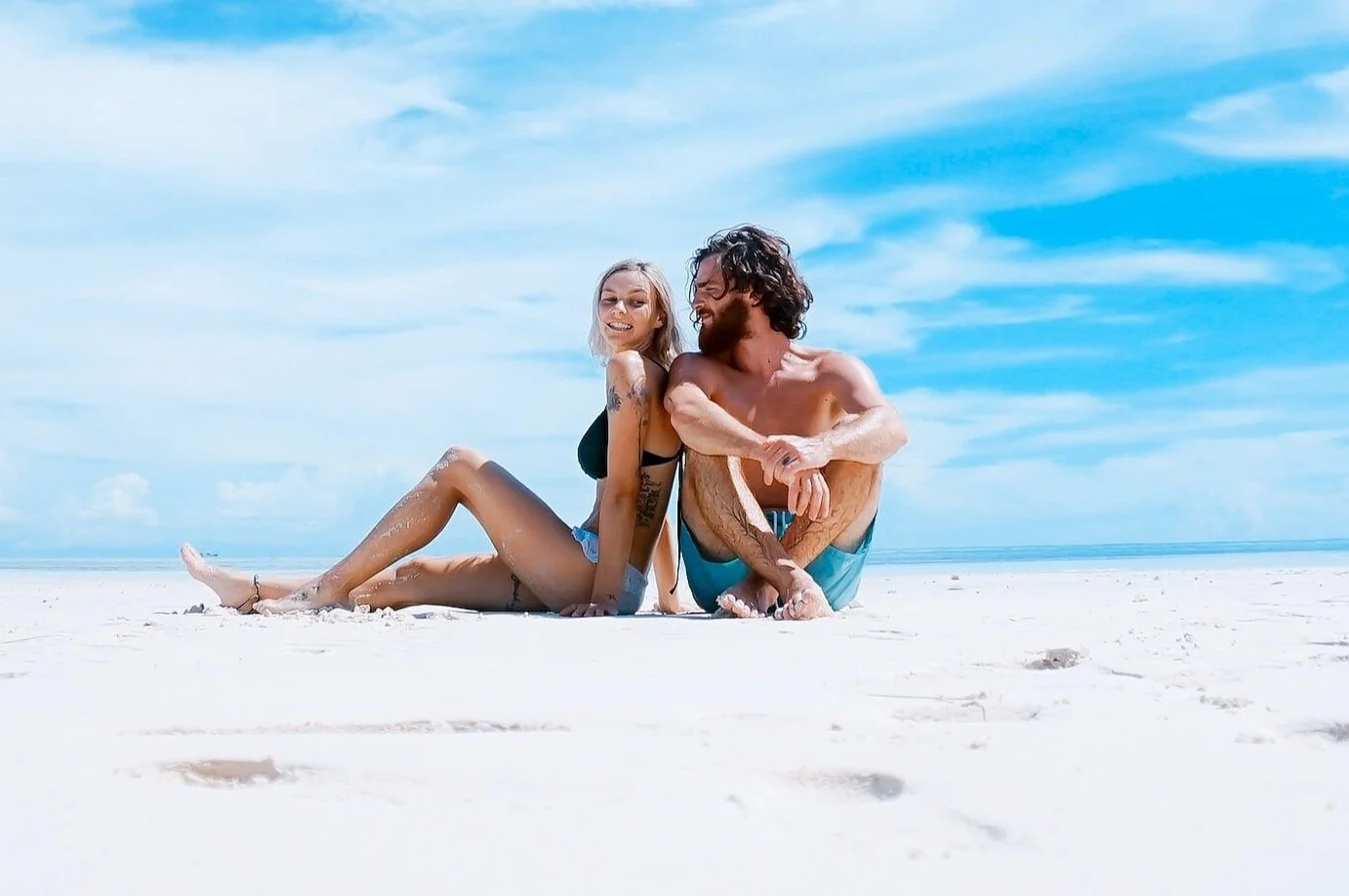 Couple sitting on a sandy beach under a blue sky, smiling and relaxing in swimsuits.