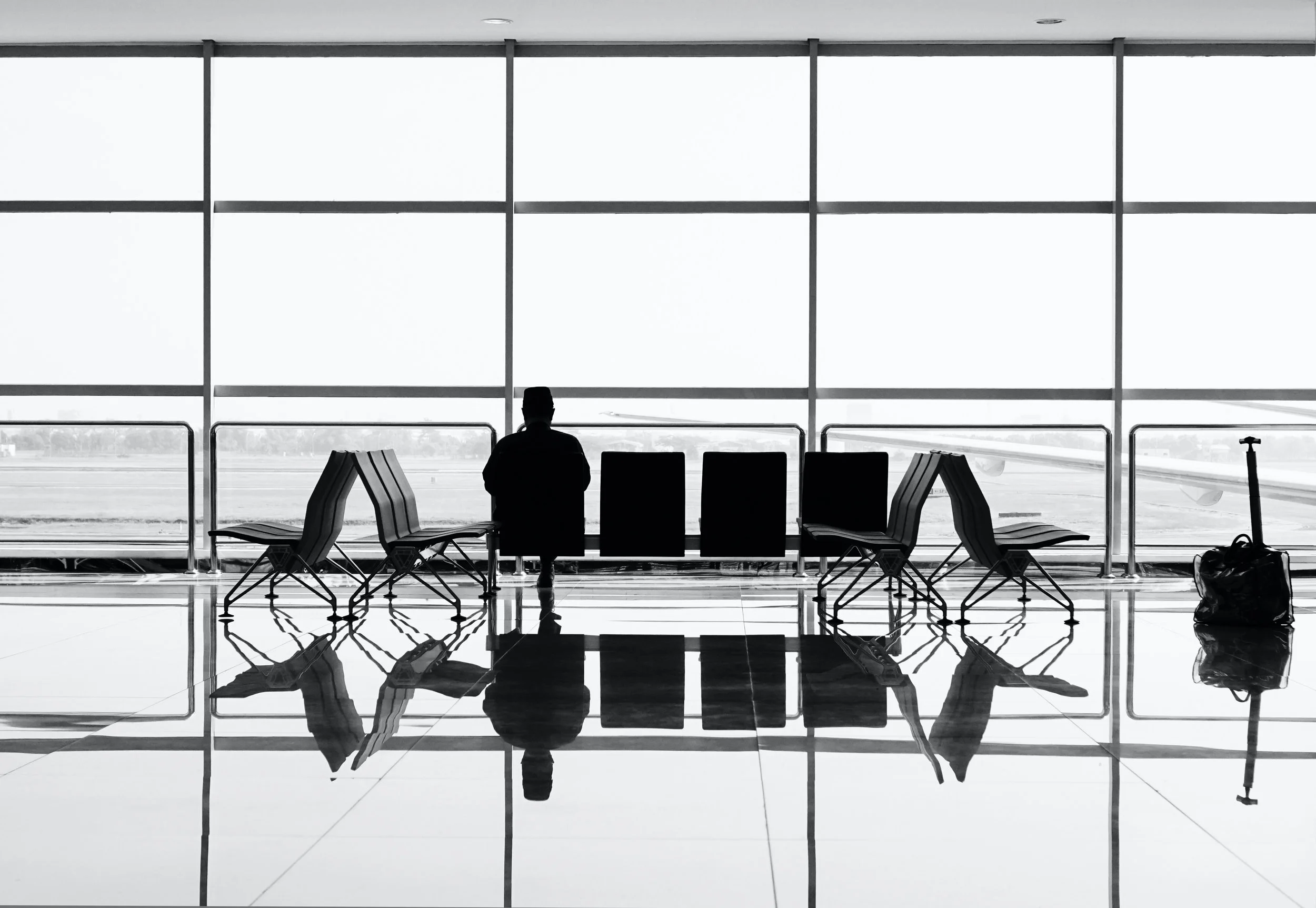 Silhouette of a person sitting in an airport waiting area with chairs, a suitcase, and large windows showing the tarmac outside.