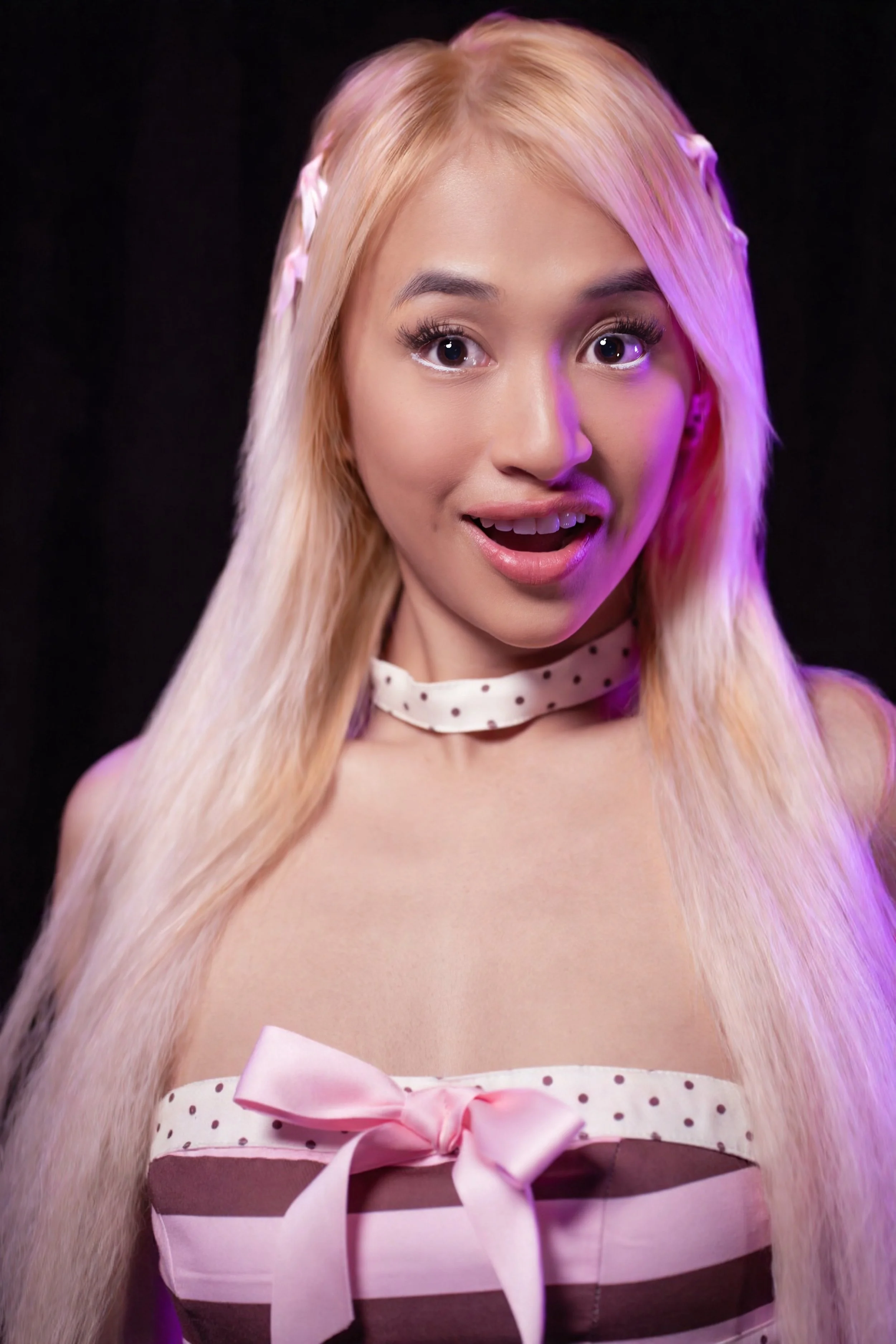 TREZIE with long blonde hair dressed in a polka dot and striped dress with a pink bow, smiling and looking surprised, against a dark background. On her first photoshoot with 72 Entertainment Group